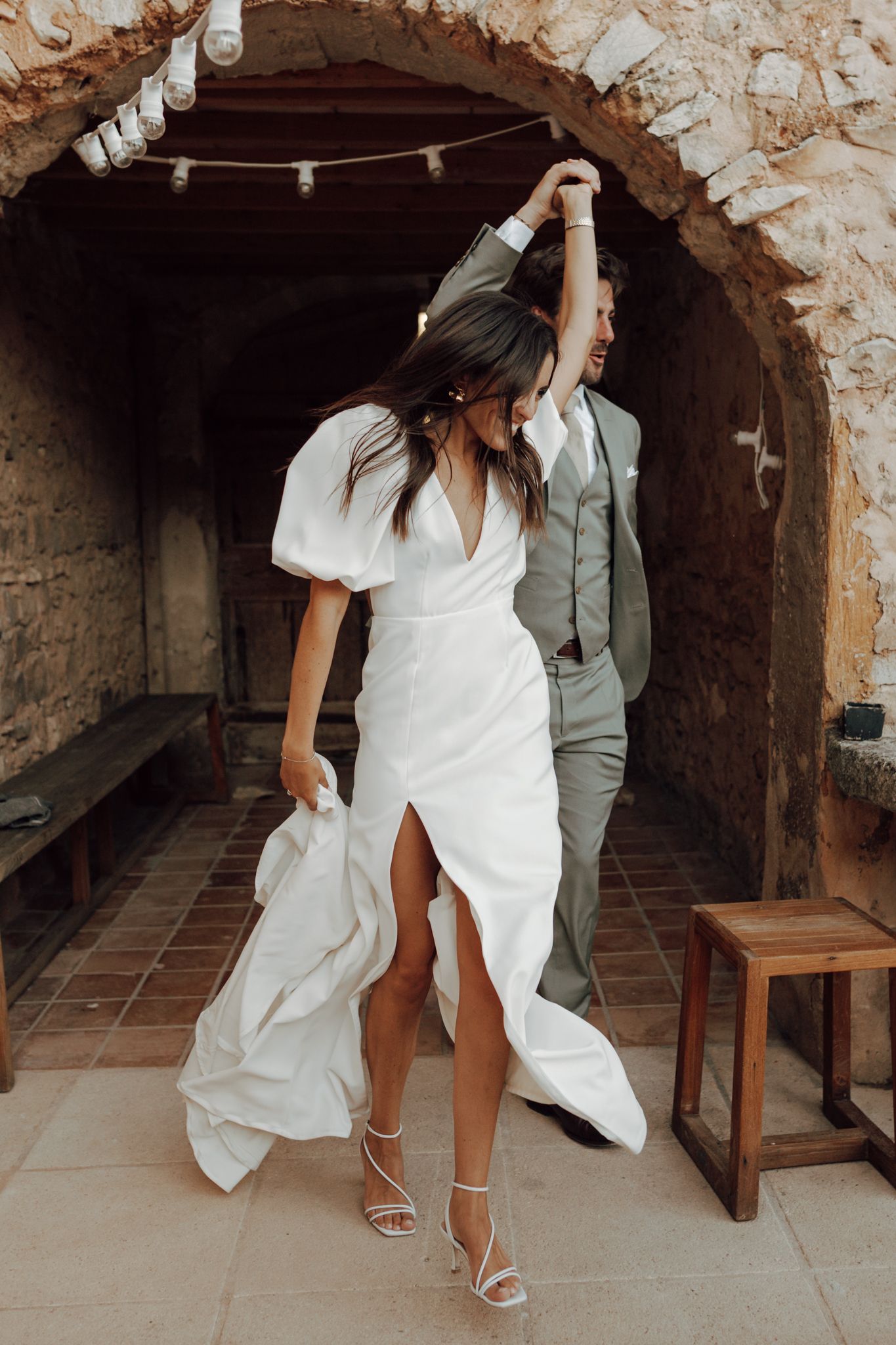Bride in white gown with groom in sage suit dancing through stone archway at elegant wedding venue.
