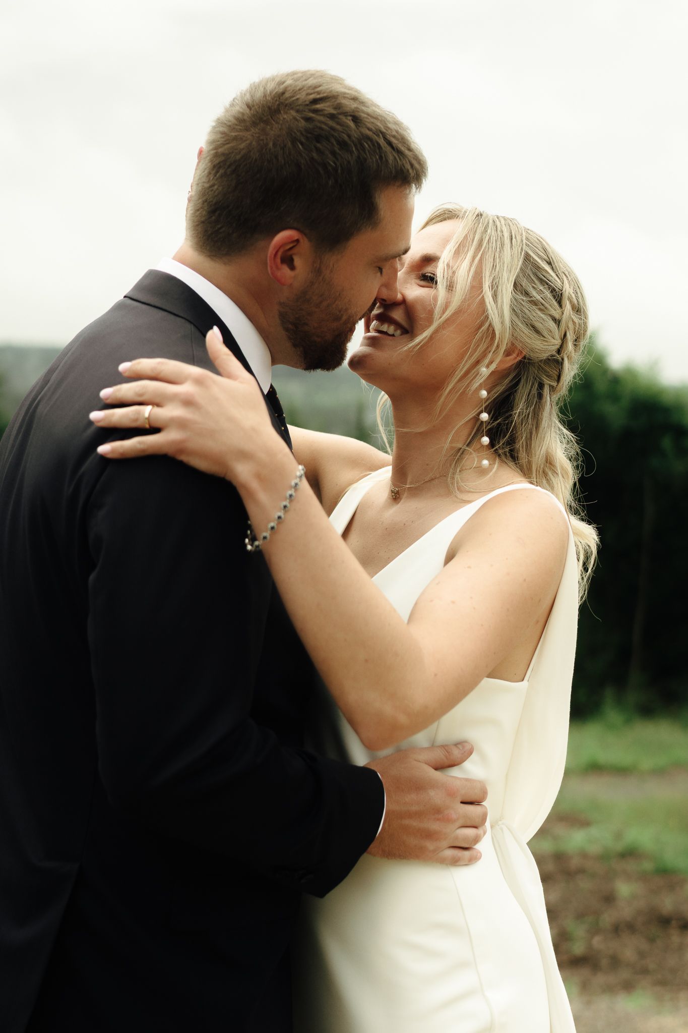 Smiling bride in white gown embraces bearded groom in black tuxedo outdoors at wedding.