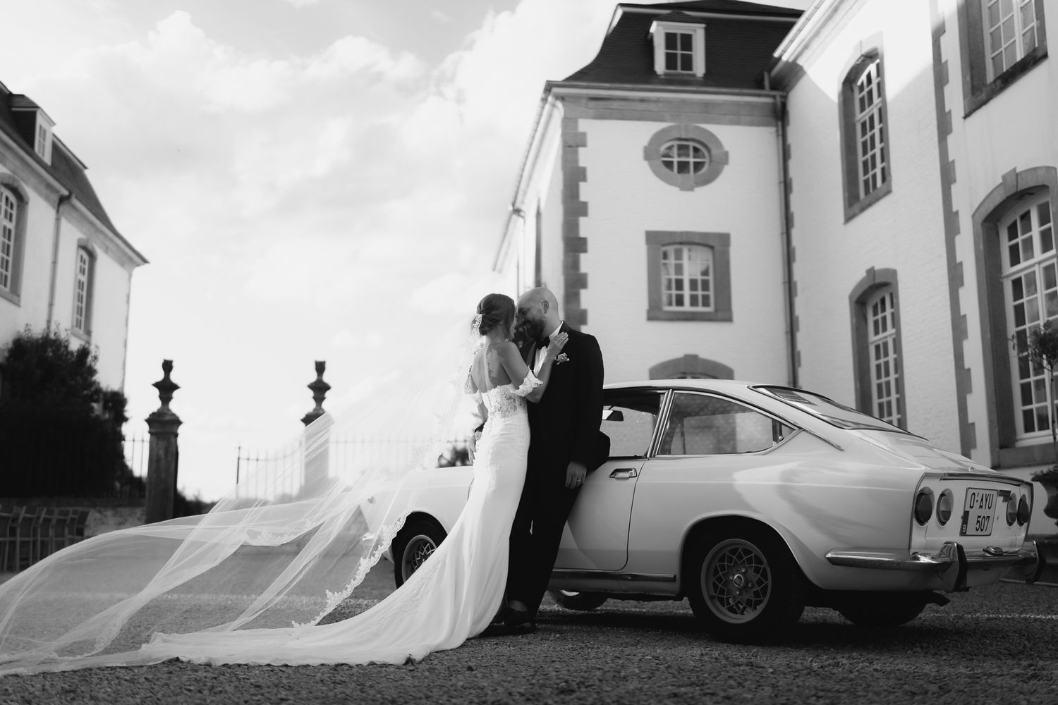 Black and white photo of bride and groom kissing beside vintage car with flowing veil at chateau venue.
