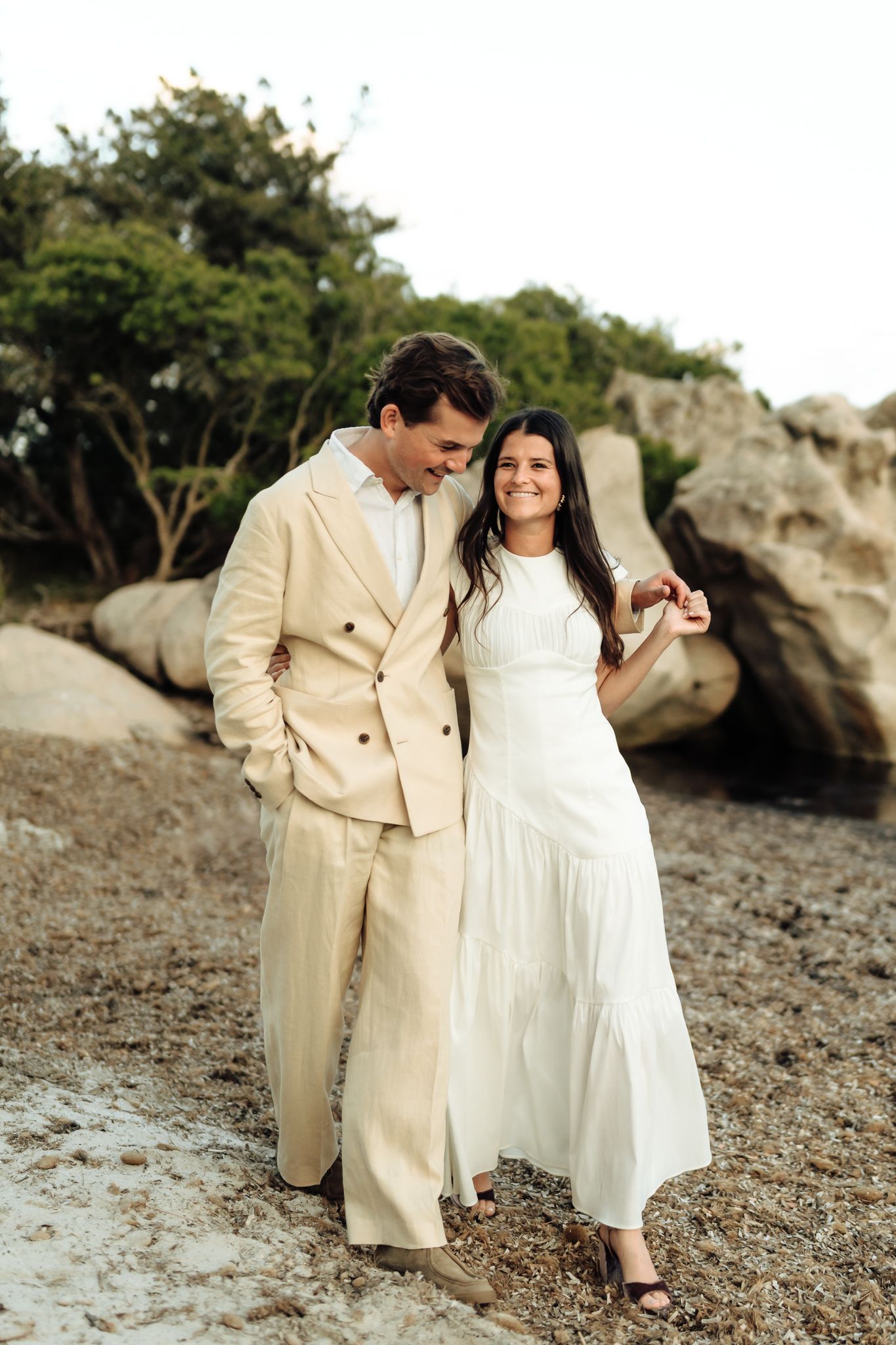Couple in matching white and beige outfits posing on a rocky beach with lush green trees behind them.