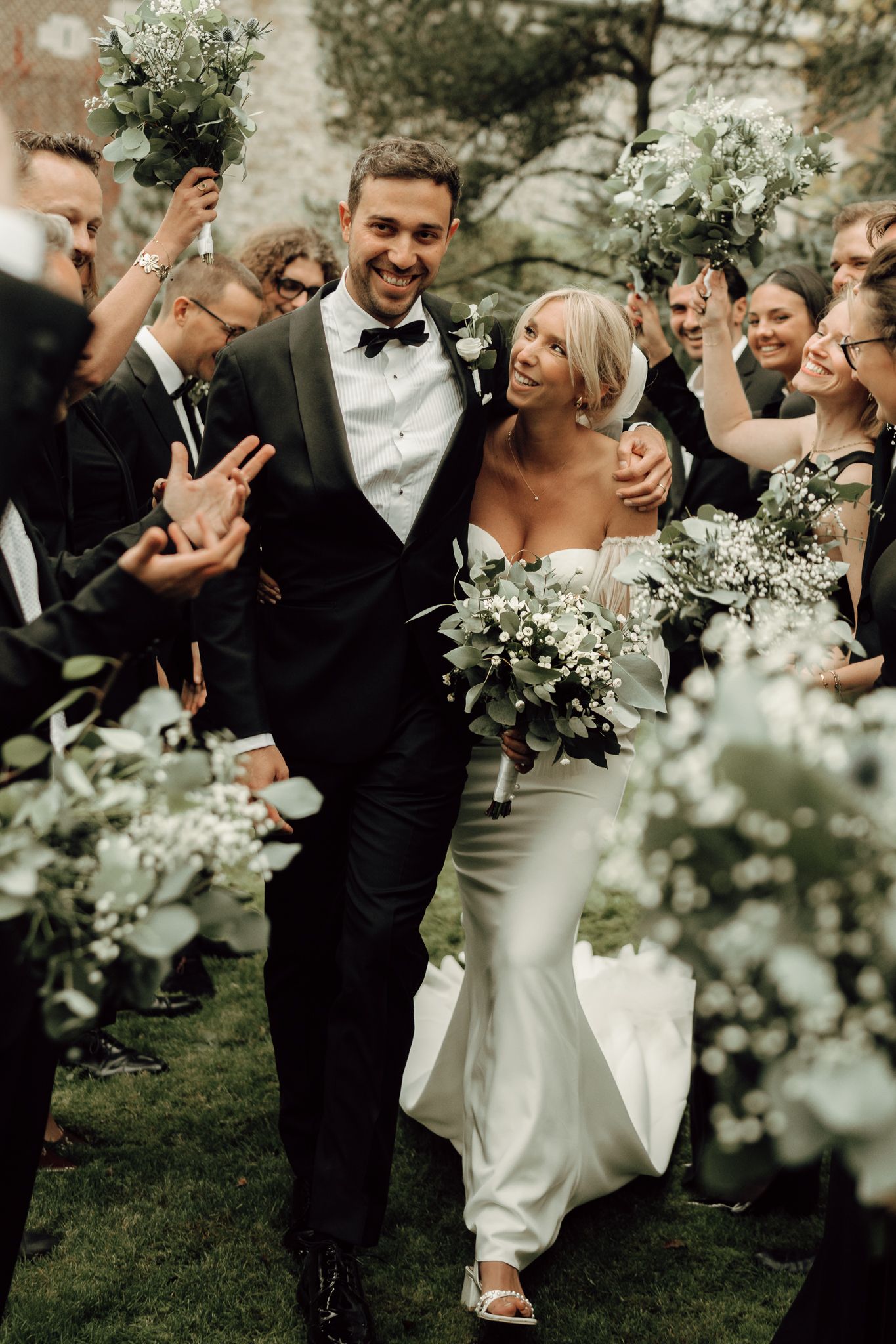 Joyful newlyweds walking through a celebratory arch of greenery bouquets held by smiling wedding guests.