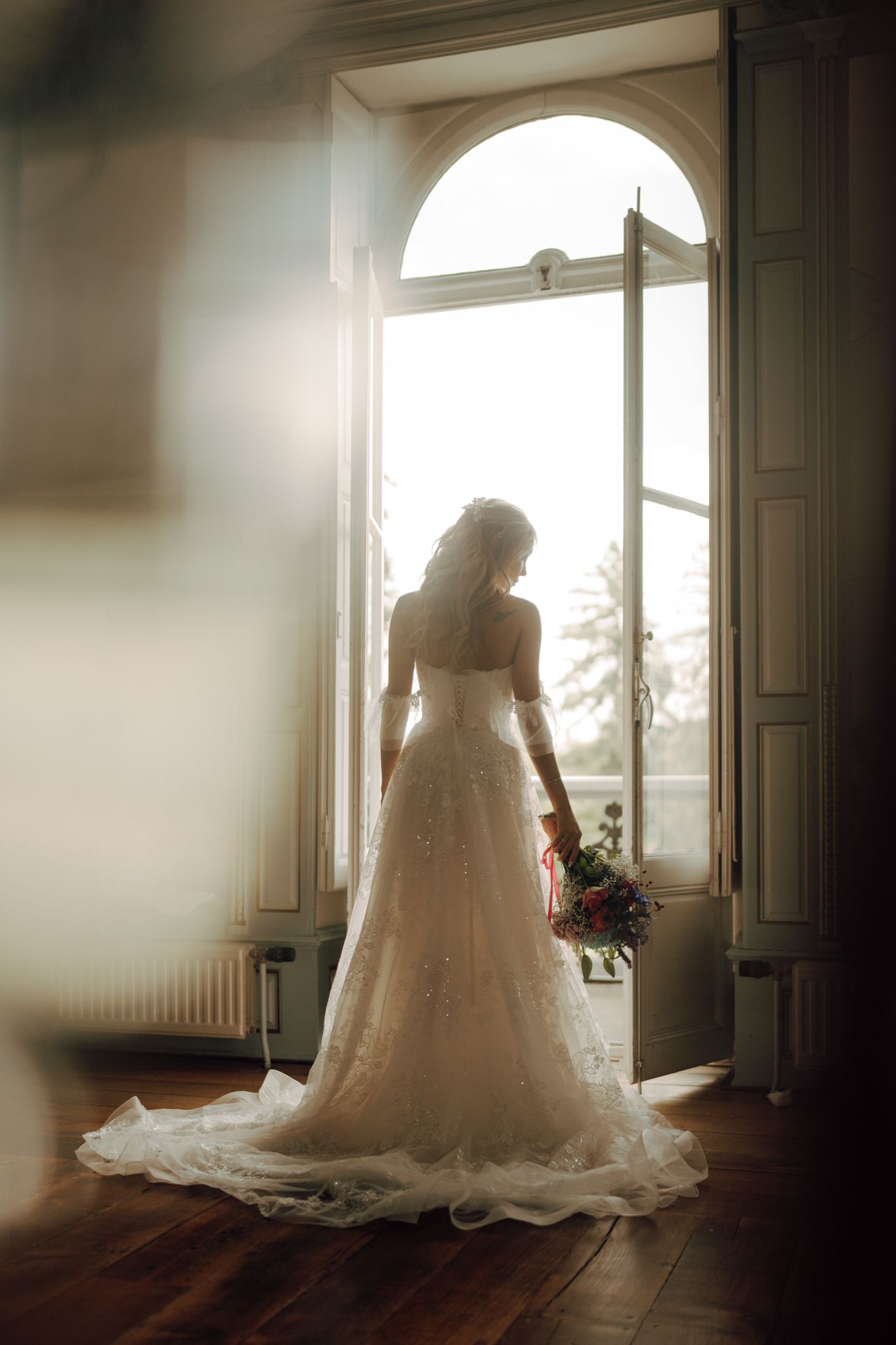 Bride in elegant lace ball gown with train standing in arched doorway with bouquet.