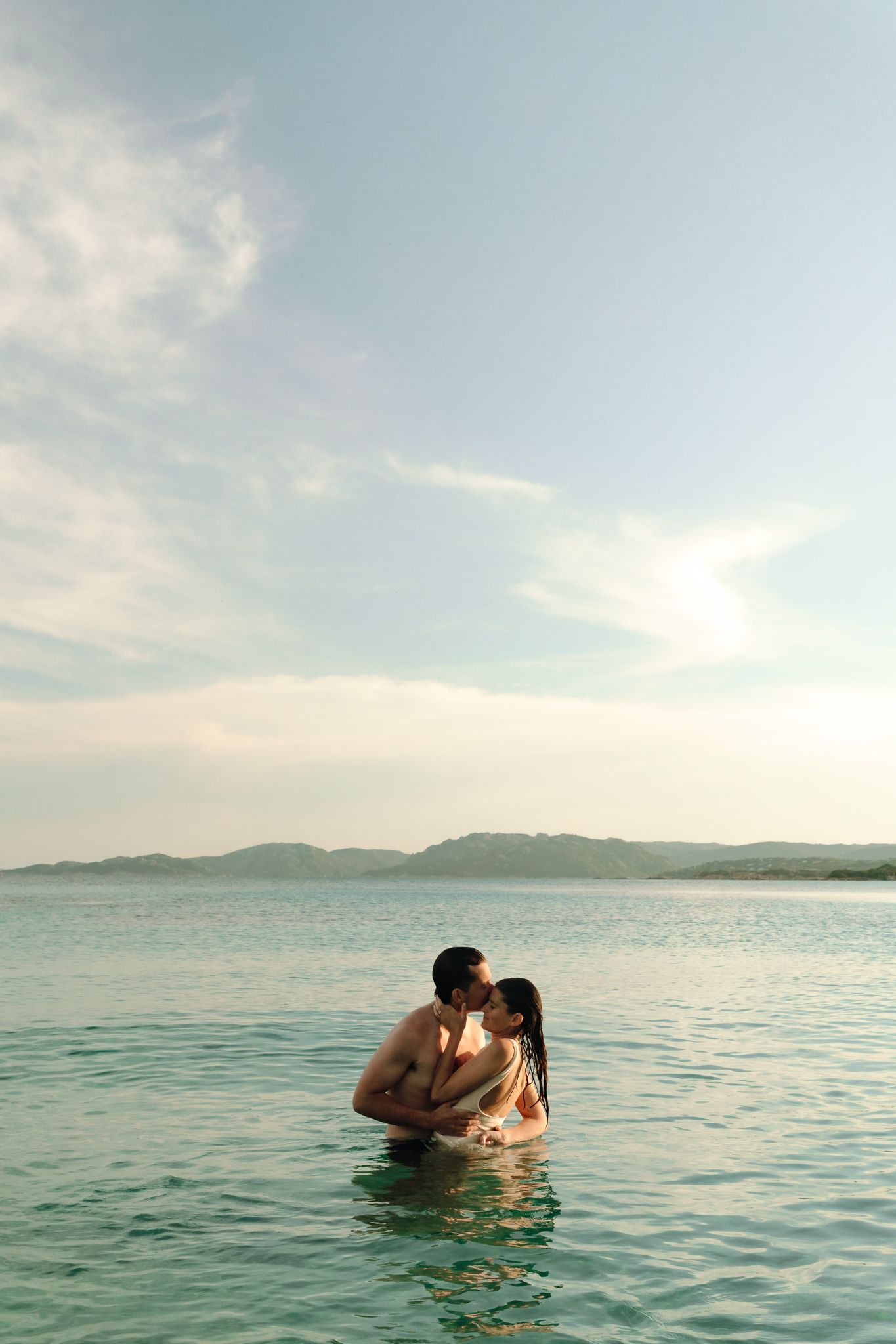 Romantic couple kissing in calm turquoise ocean water at sunset with mountains in the background.