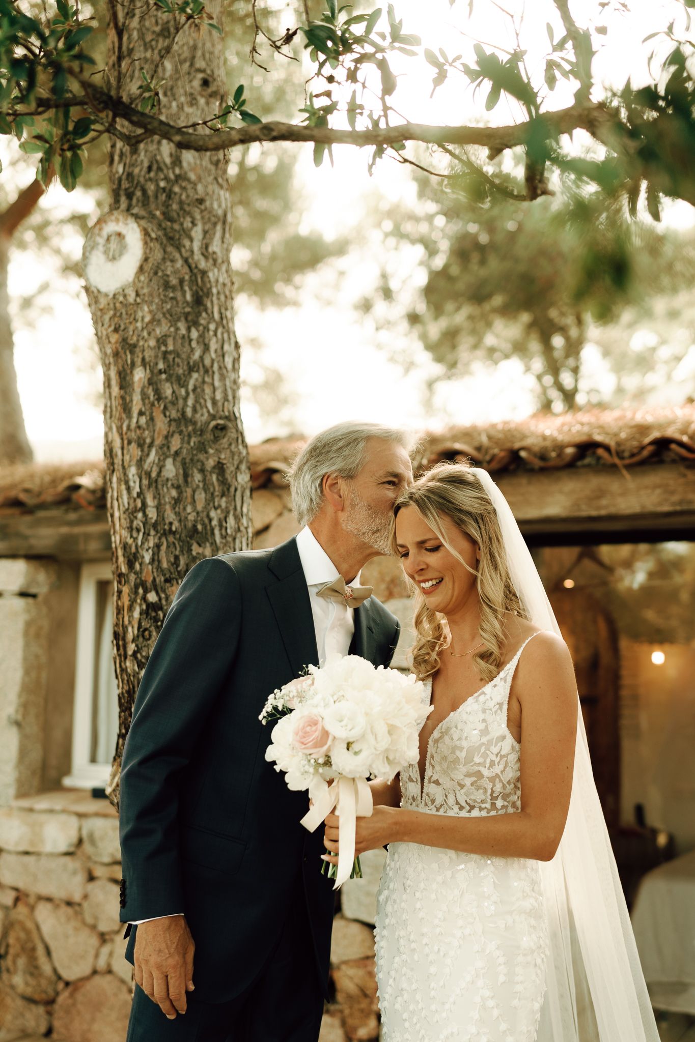 Bride and groom share tender moment holding white bouquet under olive tree outdoors.