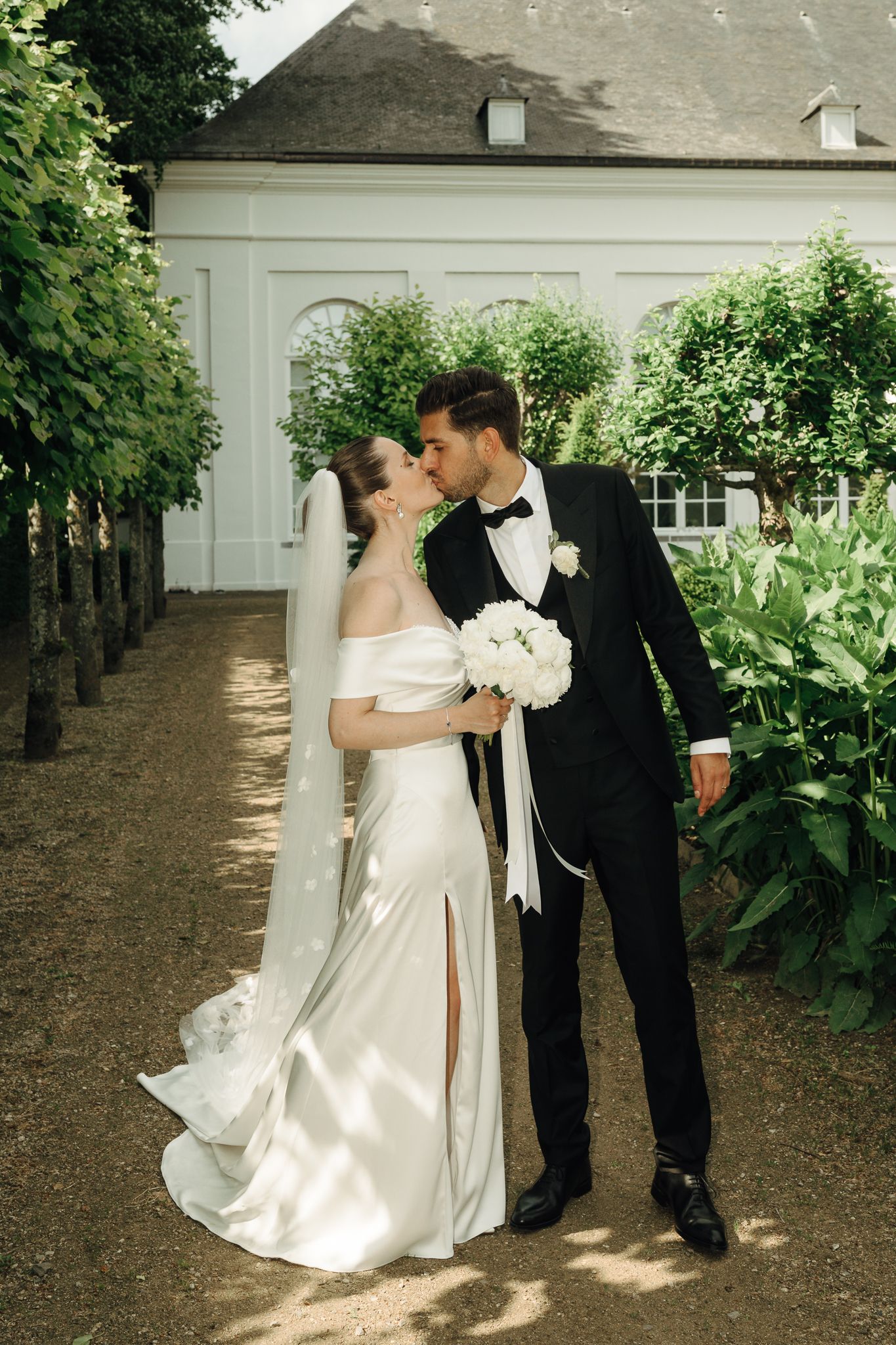 Bride and groom share a kiss on a garden path in front of a white manor surrounded by greenery.