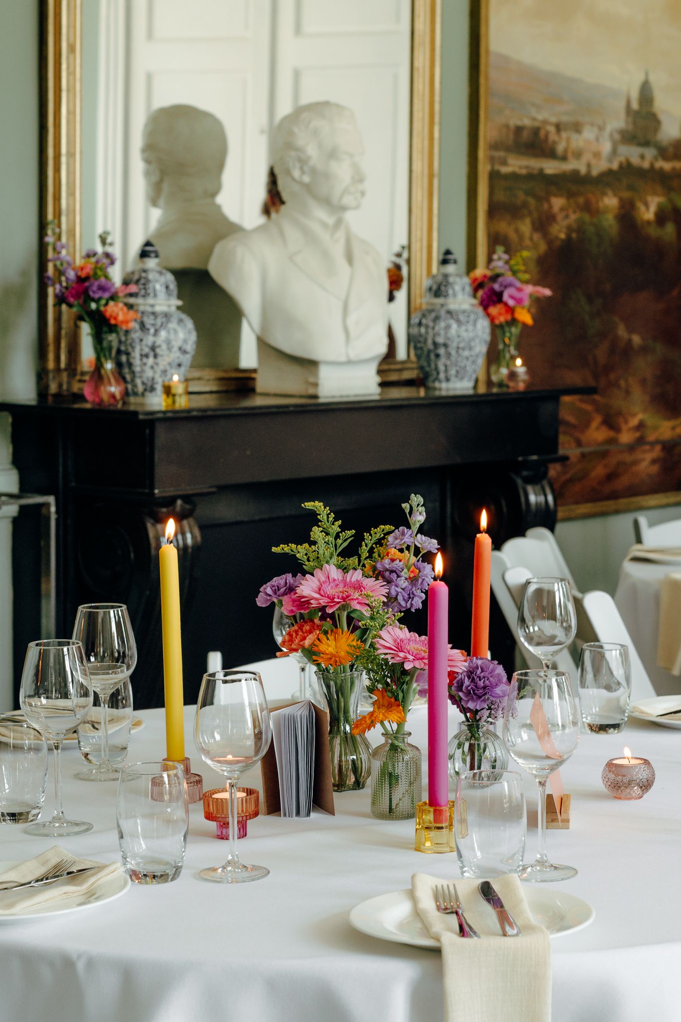 Elegant wedding reception table with colorful candles, wildflowers, crystal glasses, and classical bust sculpture.