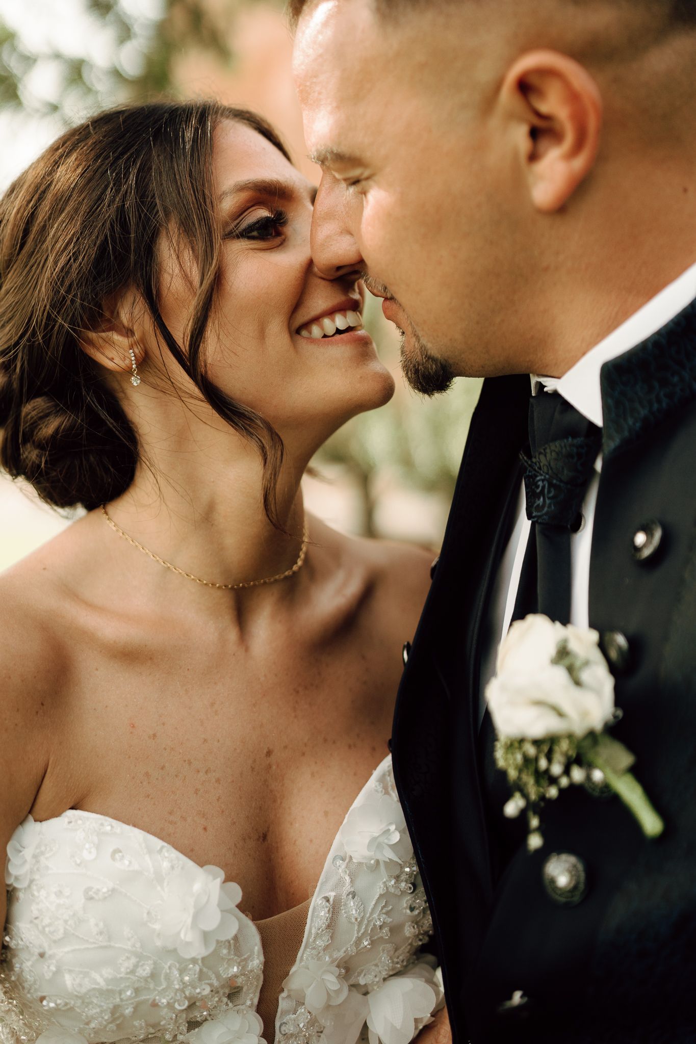 Smiling bride and groom share an intimate moment, nearly kissing, bride in white floral gown and groom in black tuxedo.