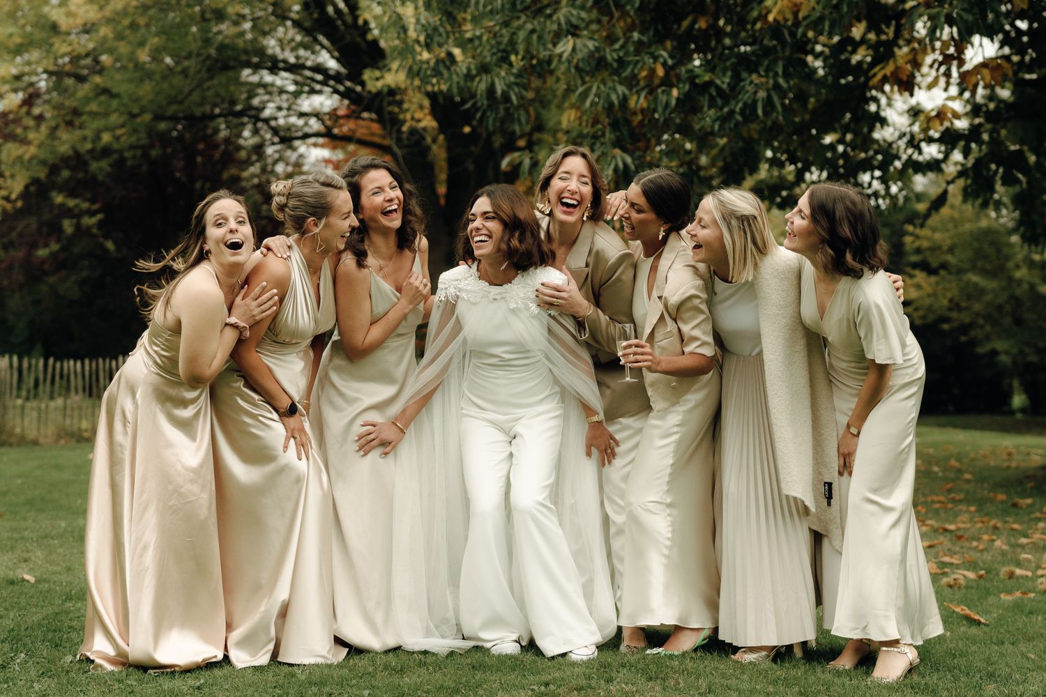 Bride laughing with bridesmaids in neutral tones on a lush green lawn at an outdoor wedding celebration.