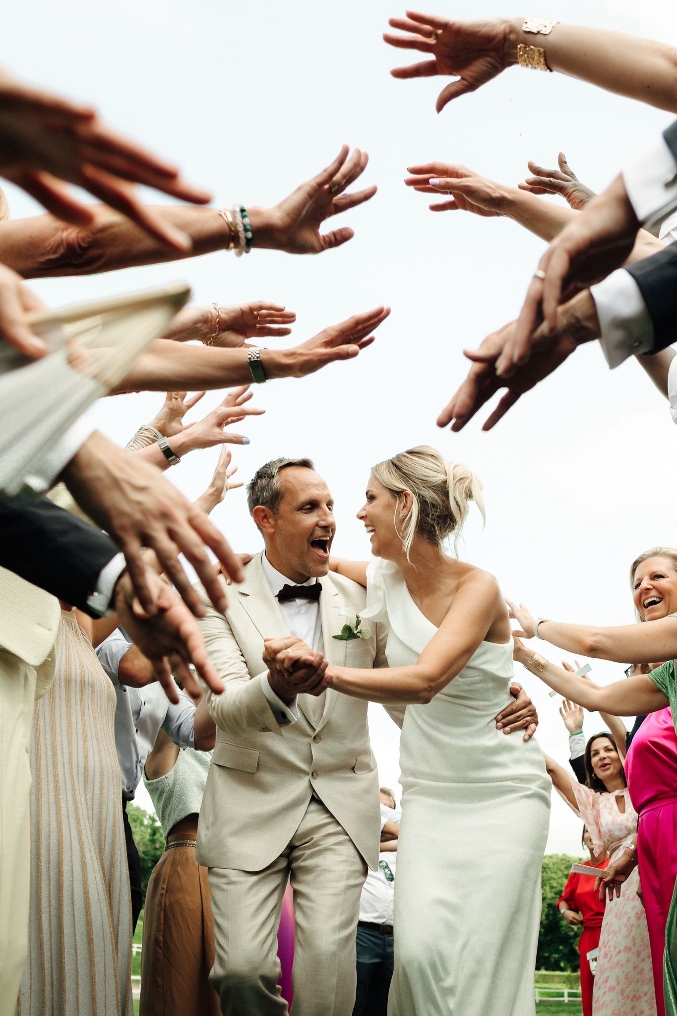 Joyful bride and groom walk through tunnel of outstretched hands from celebrating wedding guests.