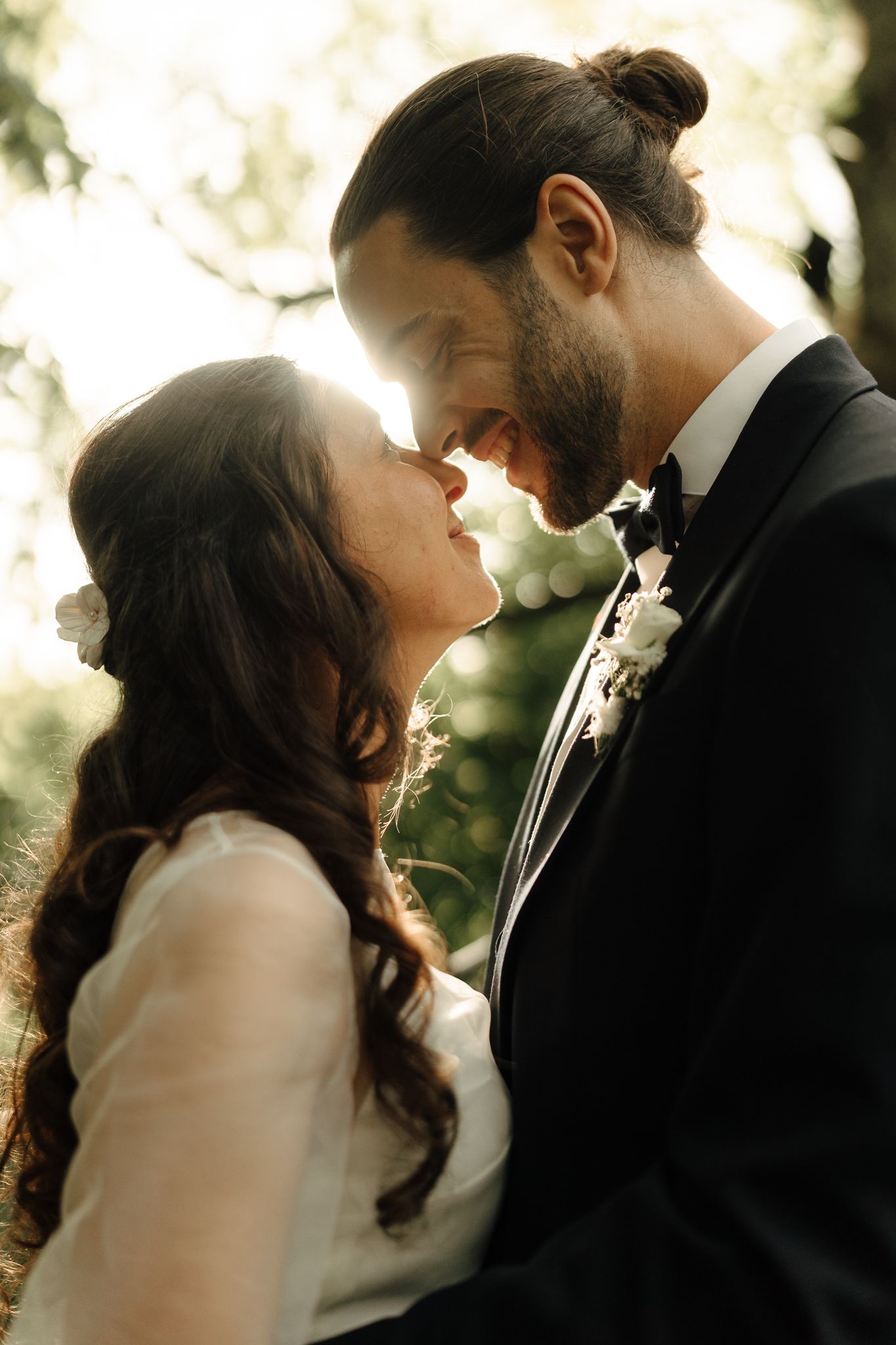 Romantic backlit portrait of bride and groom touching foreheads about to kiss in golden sunlight.