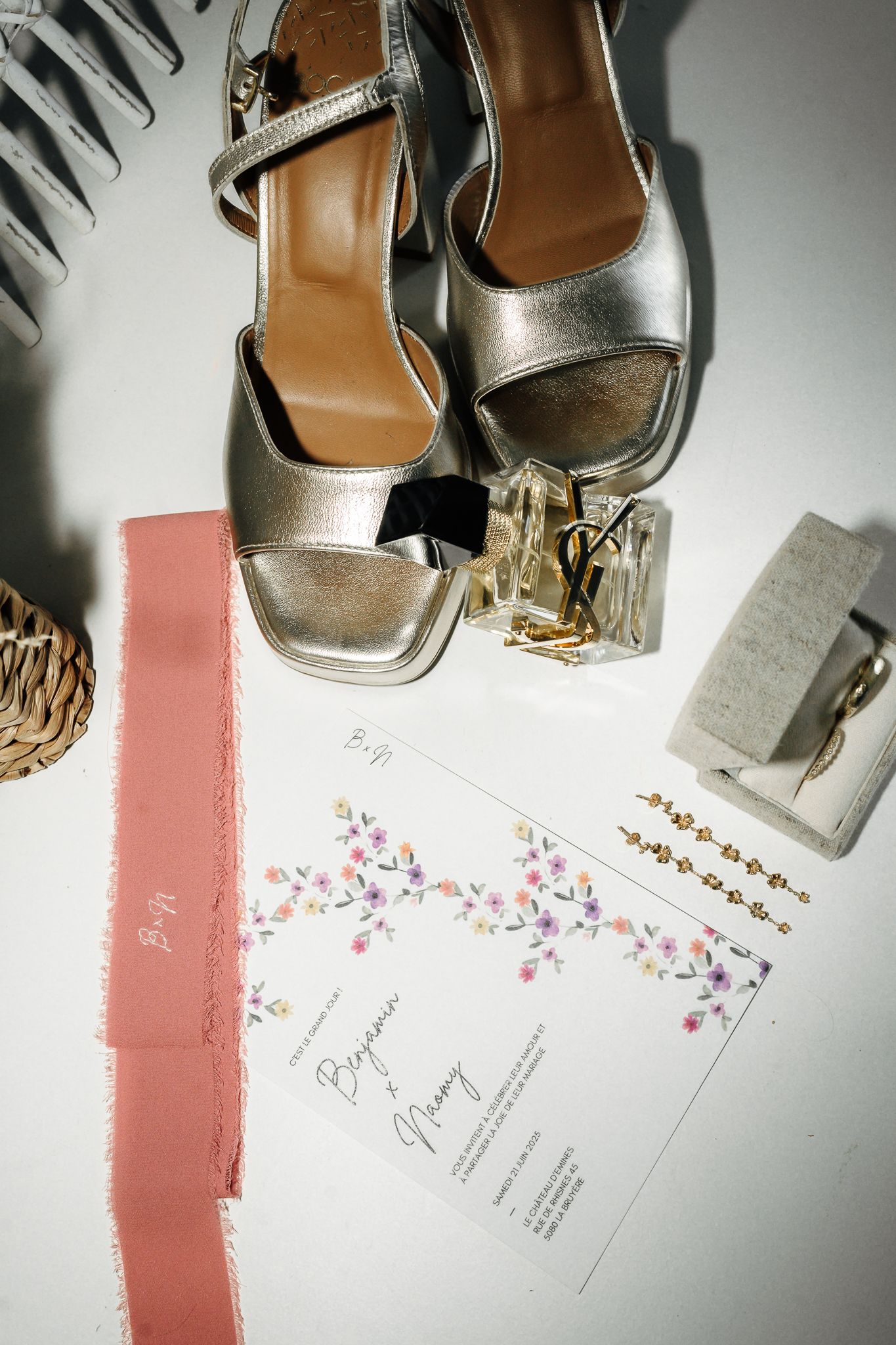 Silver strappy heels, floral wedding invitations, perfume bottle, and accessories flatlay on white surface.