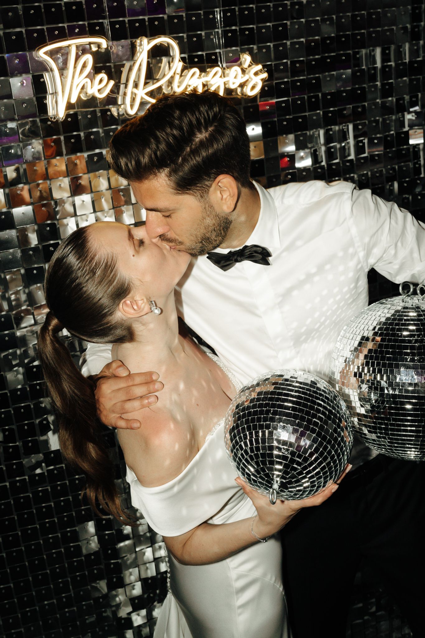Newlyweds kiss holding a disco ball in front of a mirrored wall with The Rizzos neon sign.