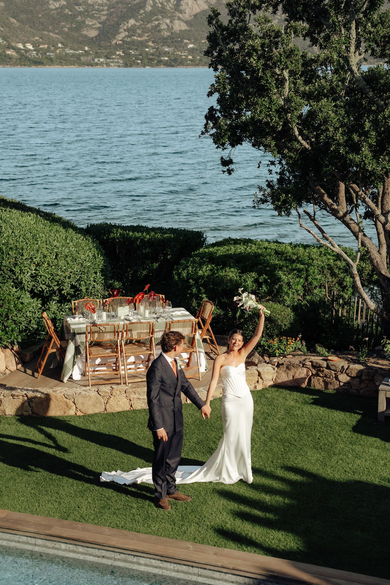 Bride and groom celebrate holding hands by a lakeside reception table on a lush green lawn with mountain views.