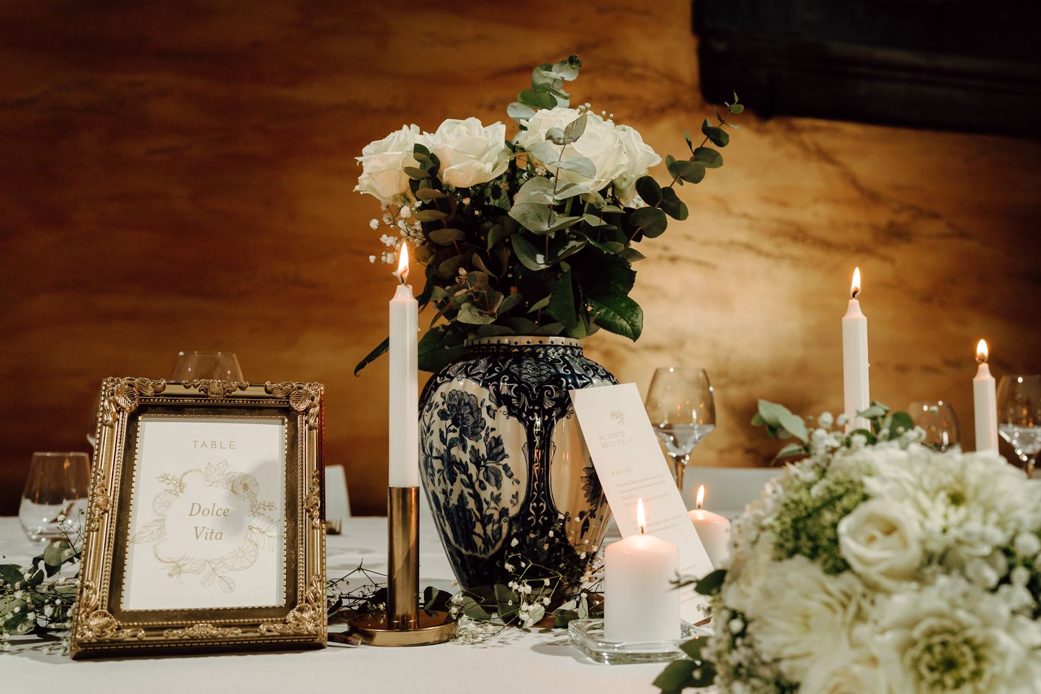 Elegant wedding reception table with white roses, candles, and gold-framed signage on a white tablecloth.