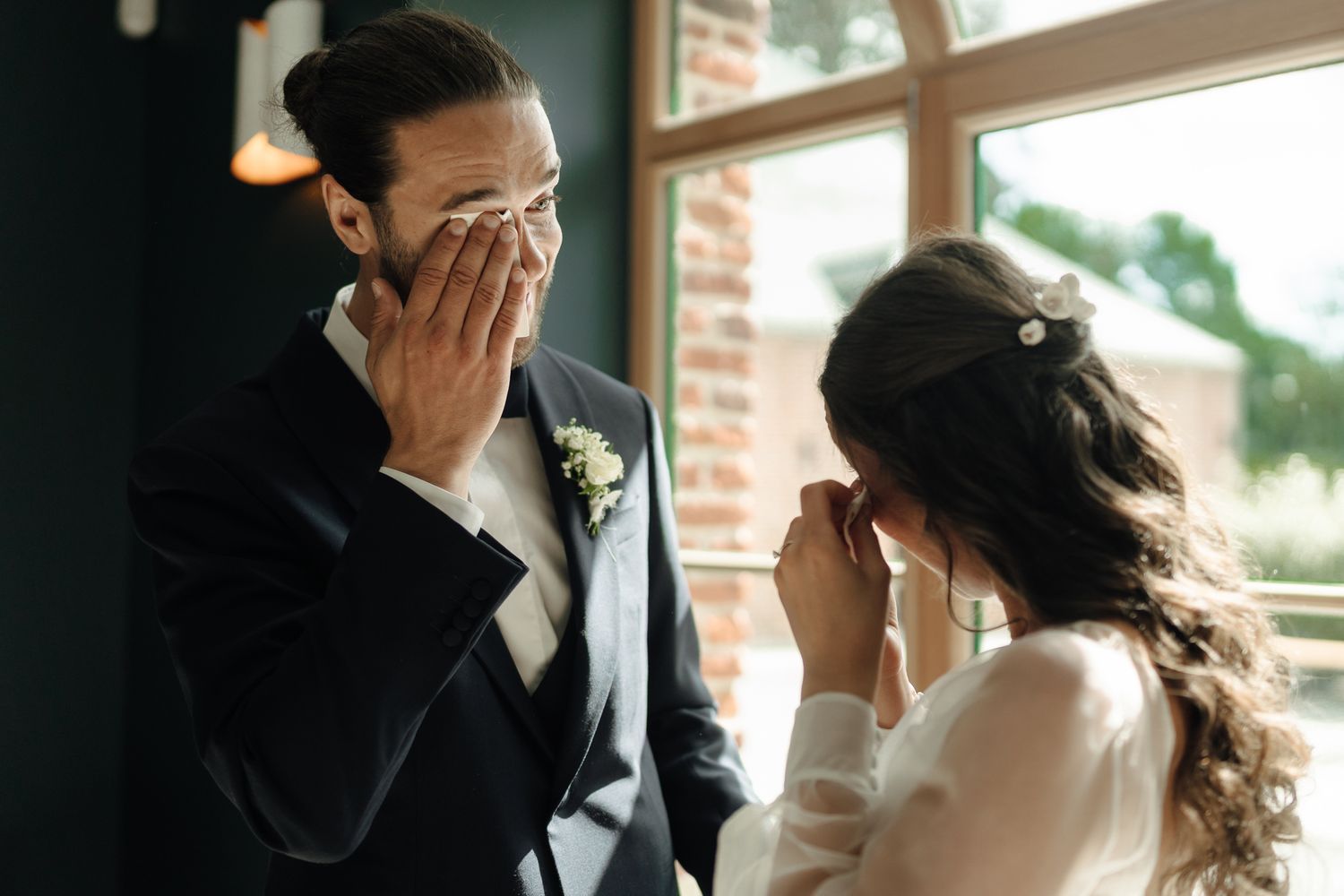 Emotional groom wiping tears while bride smiles tenderly during heartfelt first look moment indoors.