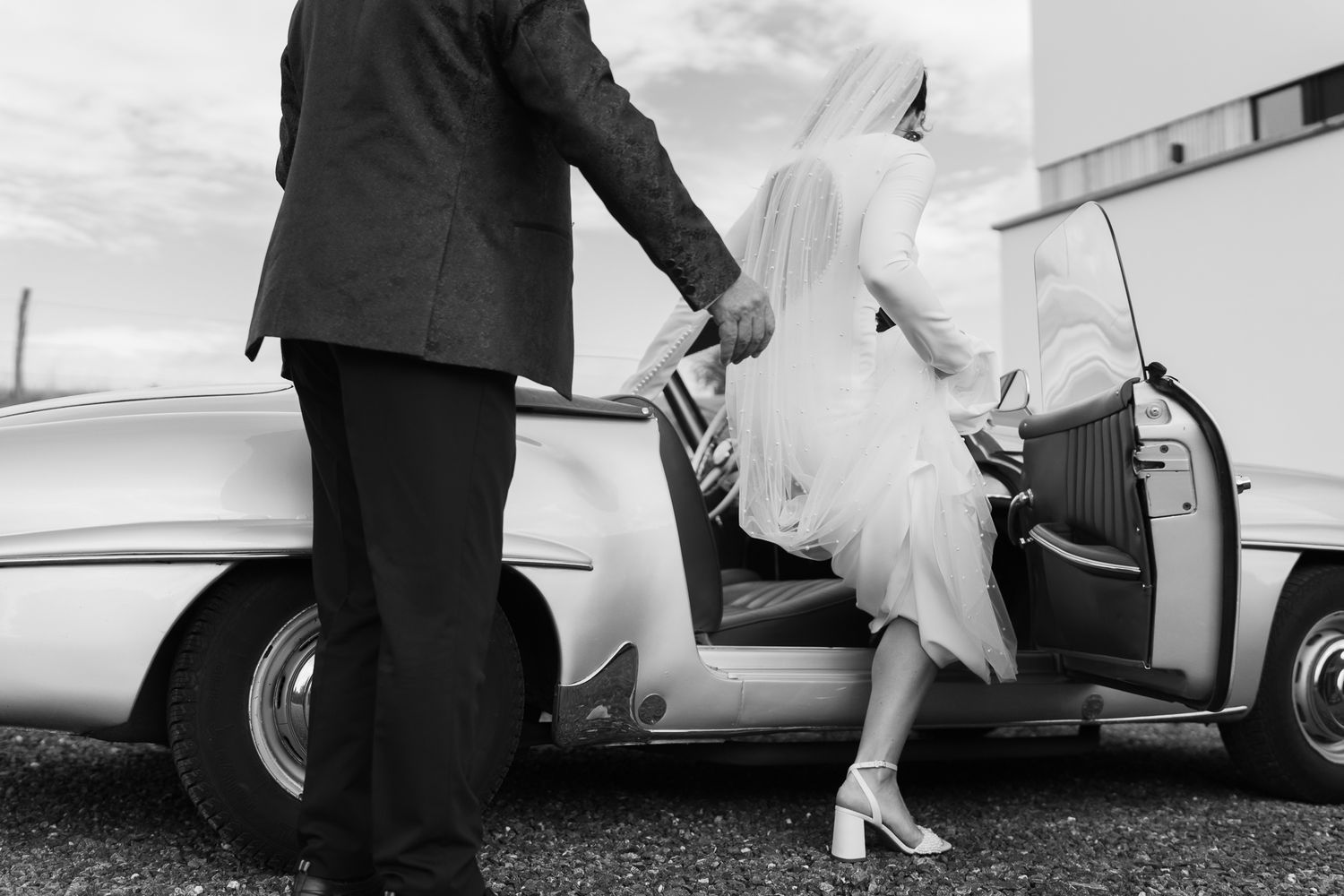 Black and white photo of groom helping bride exit vintage classic car.