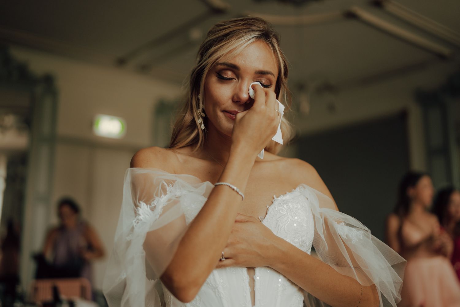 Emotional bride in white off-shoulder wedding gown wiping tears on her wedding day.