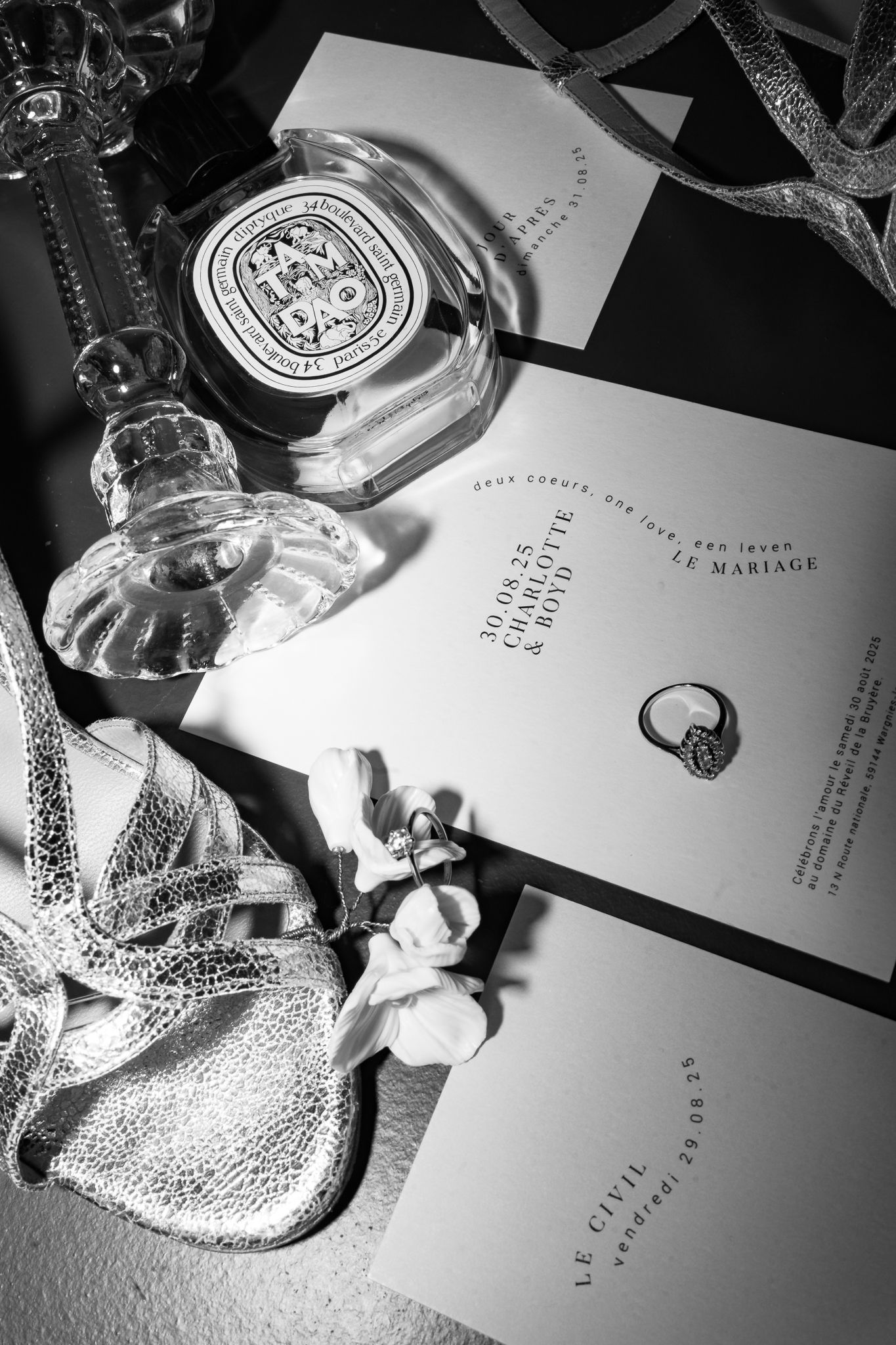 Black and white flatlay of Diptyque perfume, wedding invitations, diamond ring, and silver accessories.