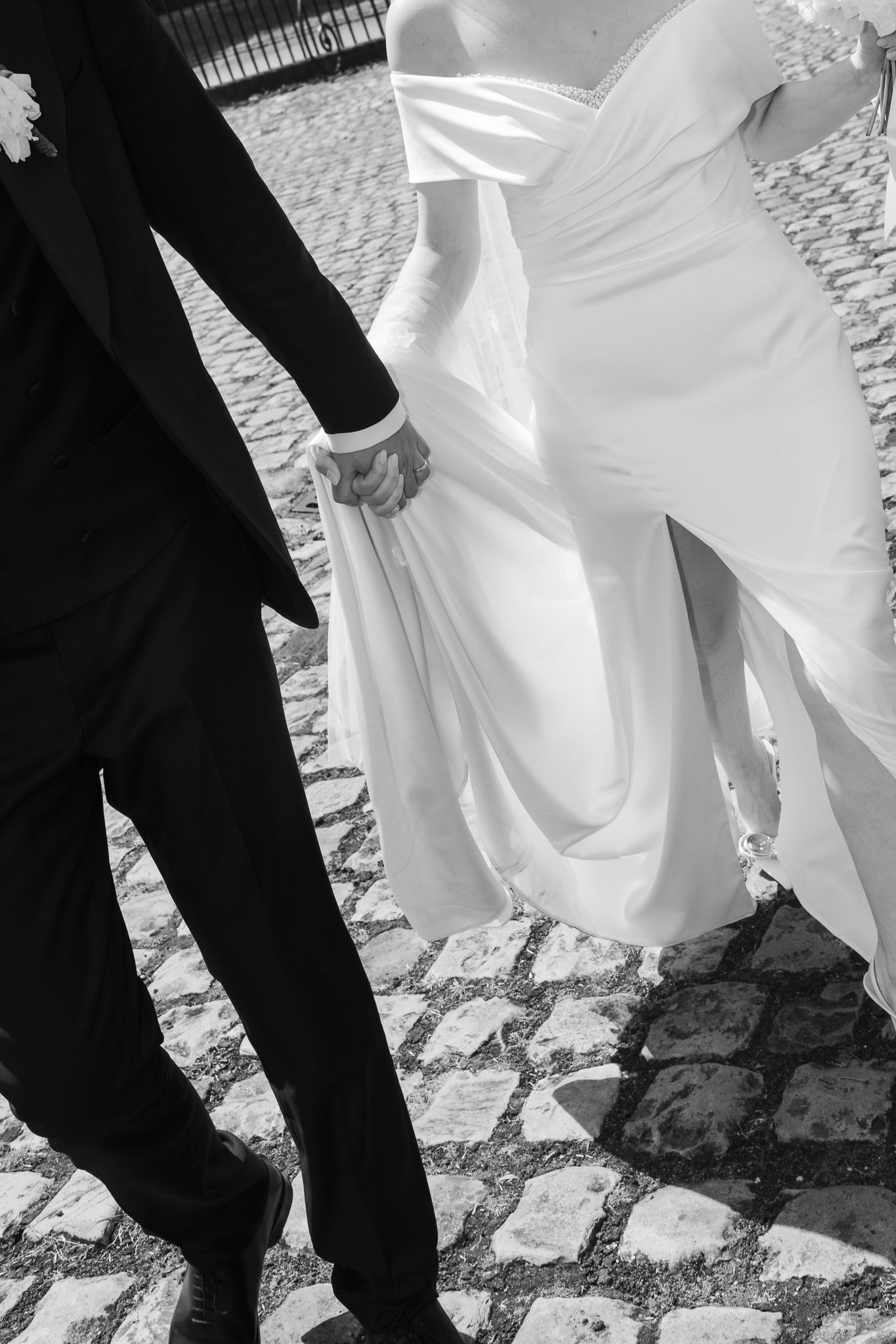 Black and white photo of bride and groom holding hands walking on cobblestone street.