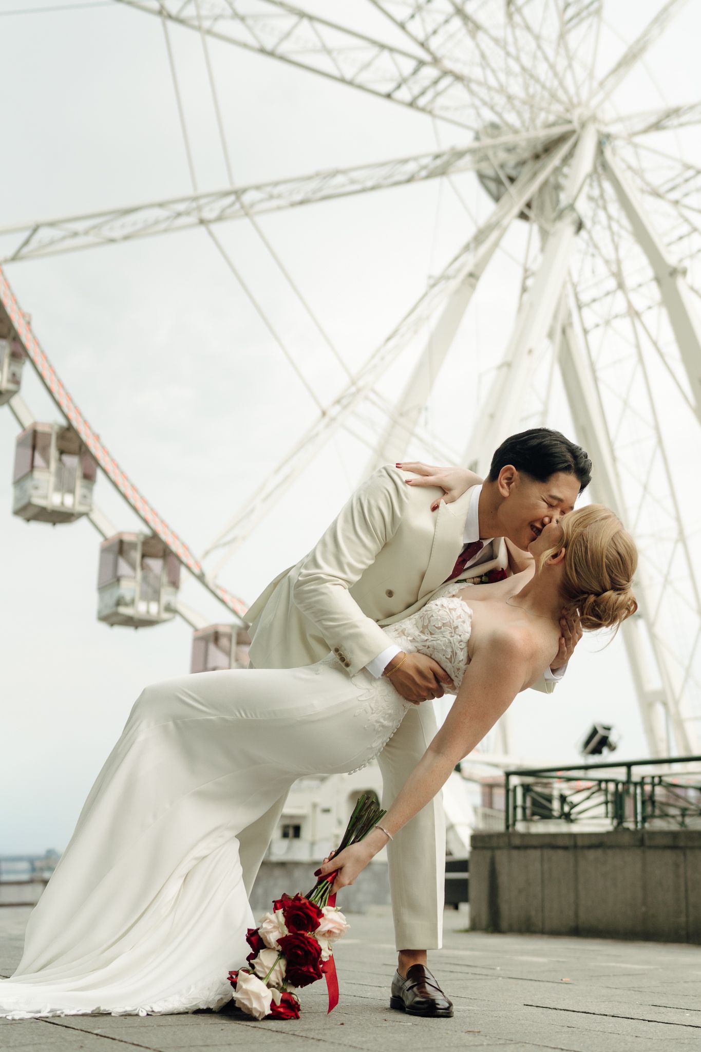 Bride and groom share a romantic dip kiss in front of a ferris wheel, bride holding red roses bouquet.