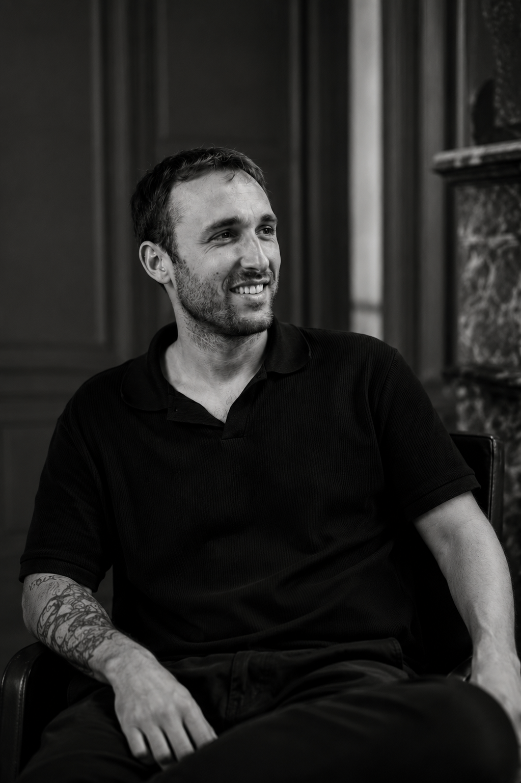 Black and white portrait of a smiling man with tattoos wearing a black polo shirt, seated in a chair.