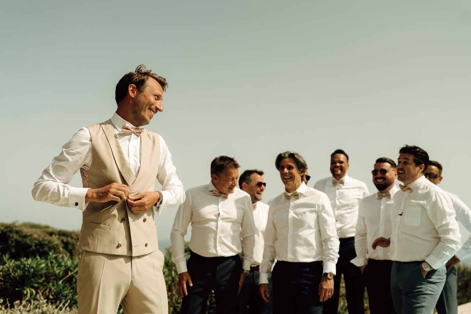 Groom in beige vest and bow tie smiling at groomsmen in white shirts on a sunny outdoor wedding.