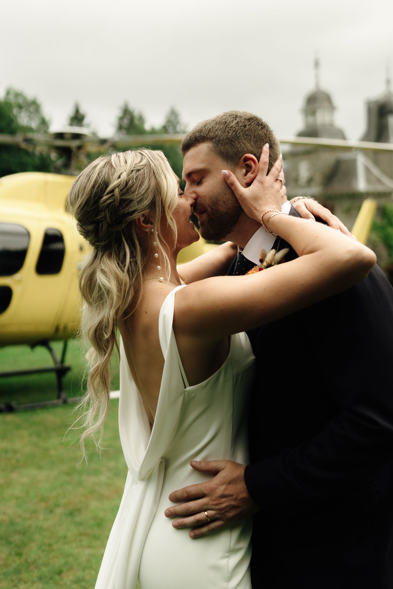 Bride and groom share a kiss in front of a yellow helicopter on a green lawn.