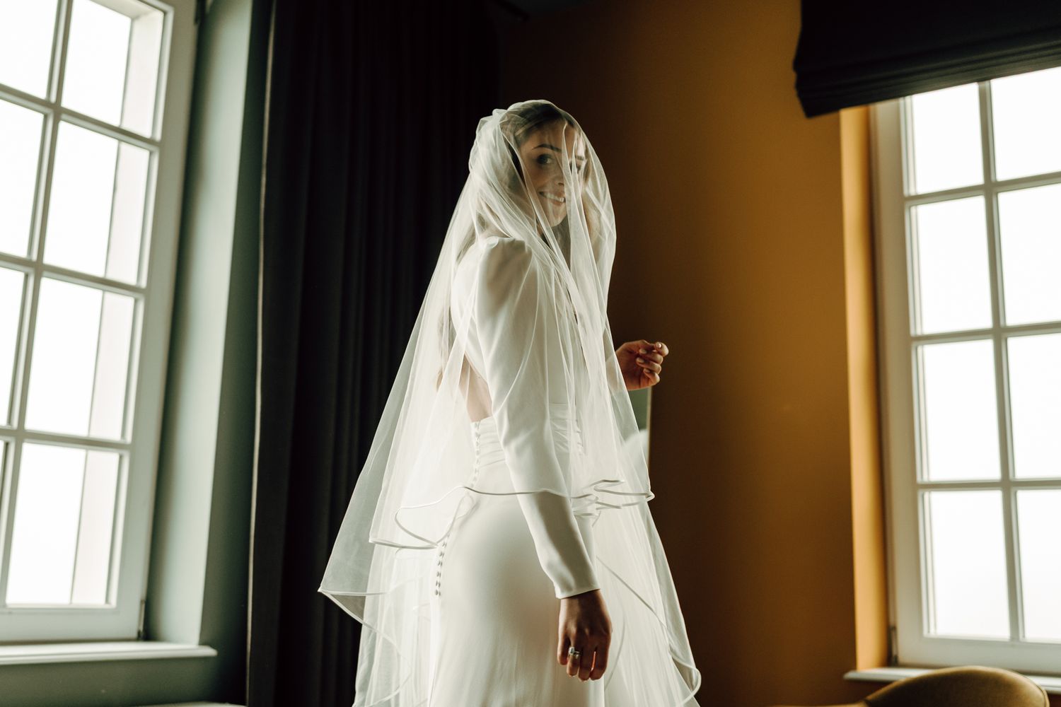 Bride in a two-tier white veil and fitted wedding dress standing between tall windows in soft light.