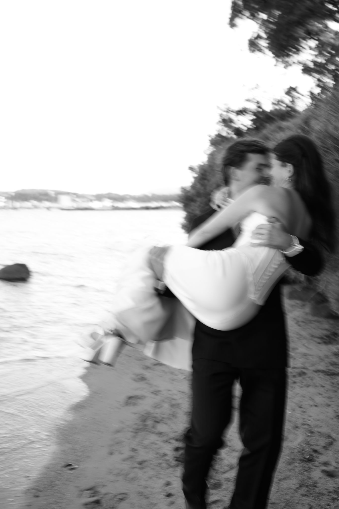 Black and white photo of a groom lifting his bride on a beach shoreline, sharing a romantic kiss.