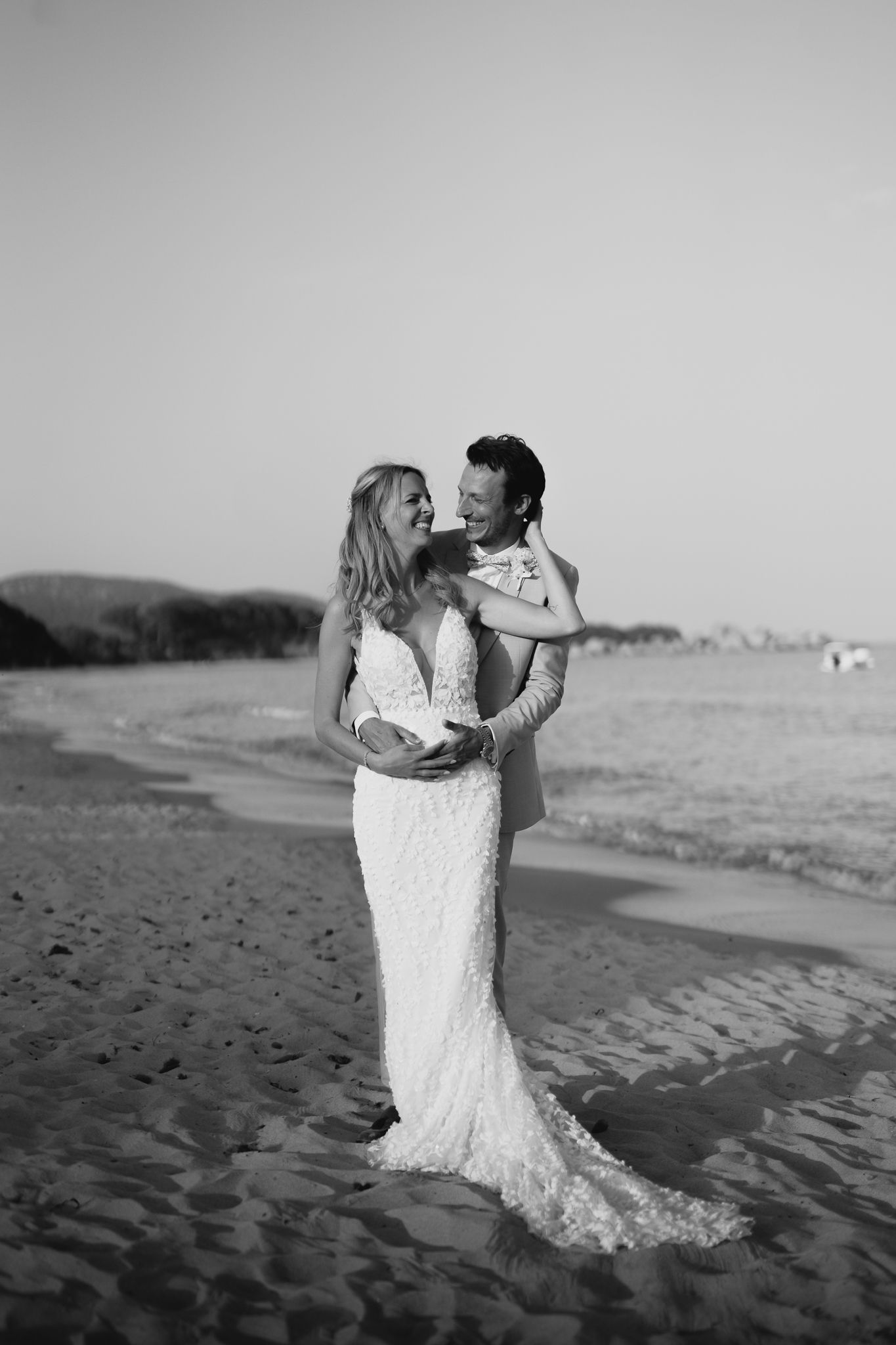 Black and white photo of bride and groom embracing joyfully on a sandy beach in wedding attire.
