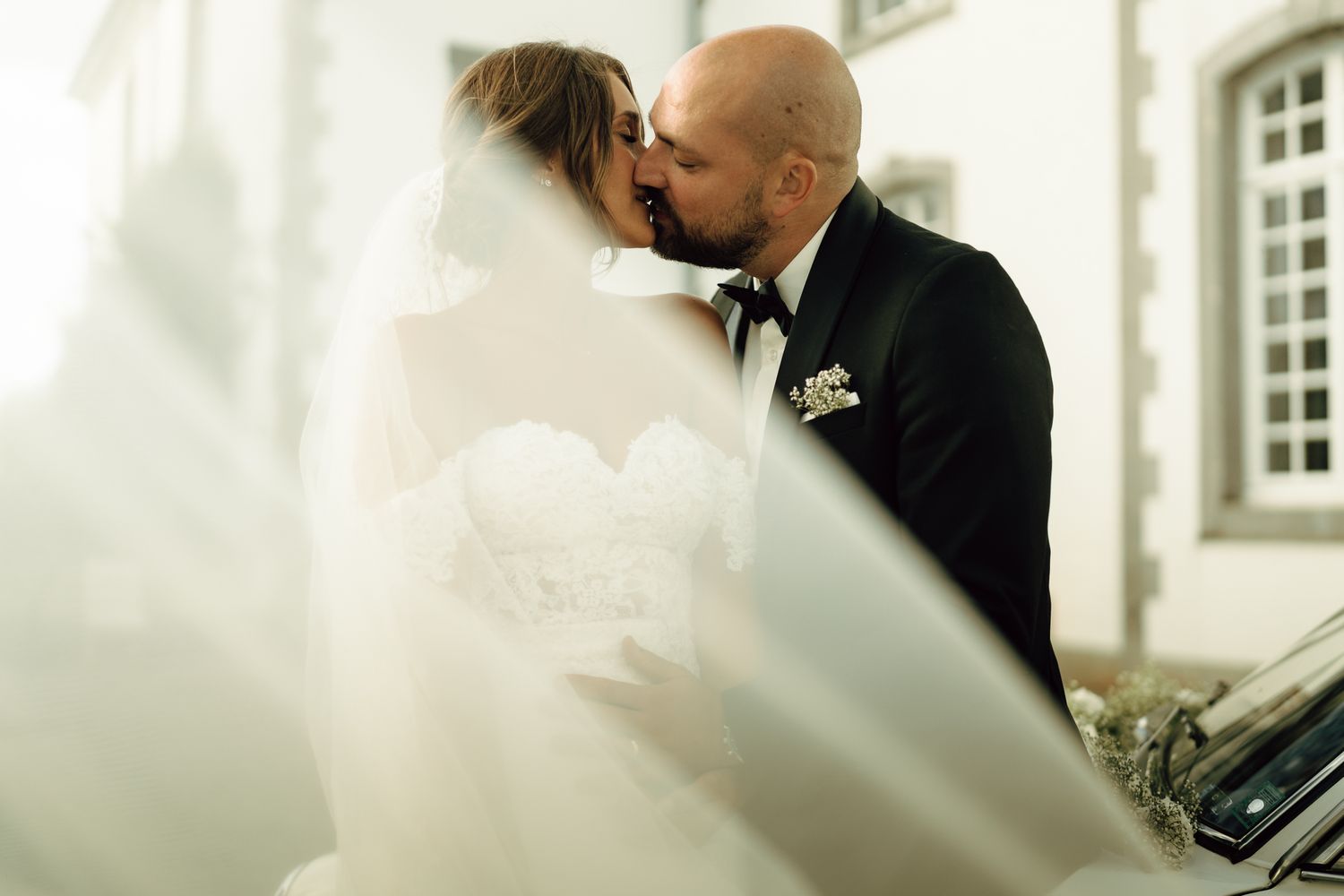 Bride and groom share a passionate kiss framed by flowing lace wedding veil outside a chateau venue.