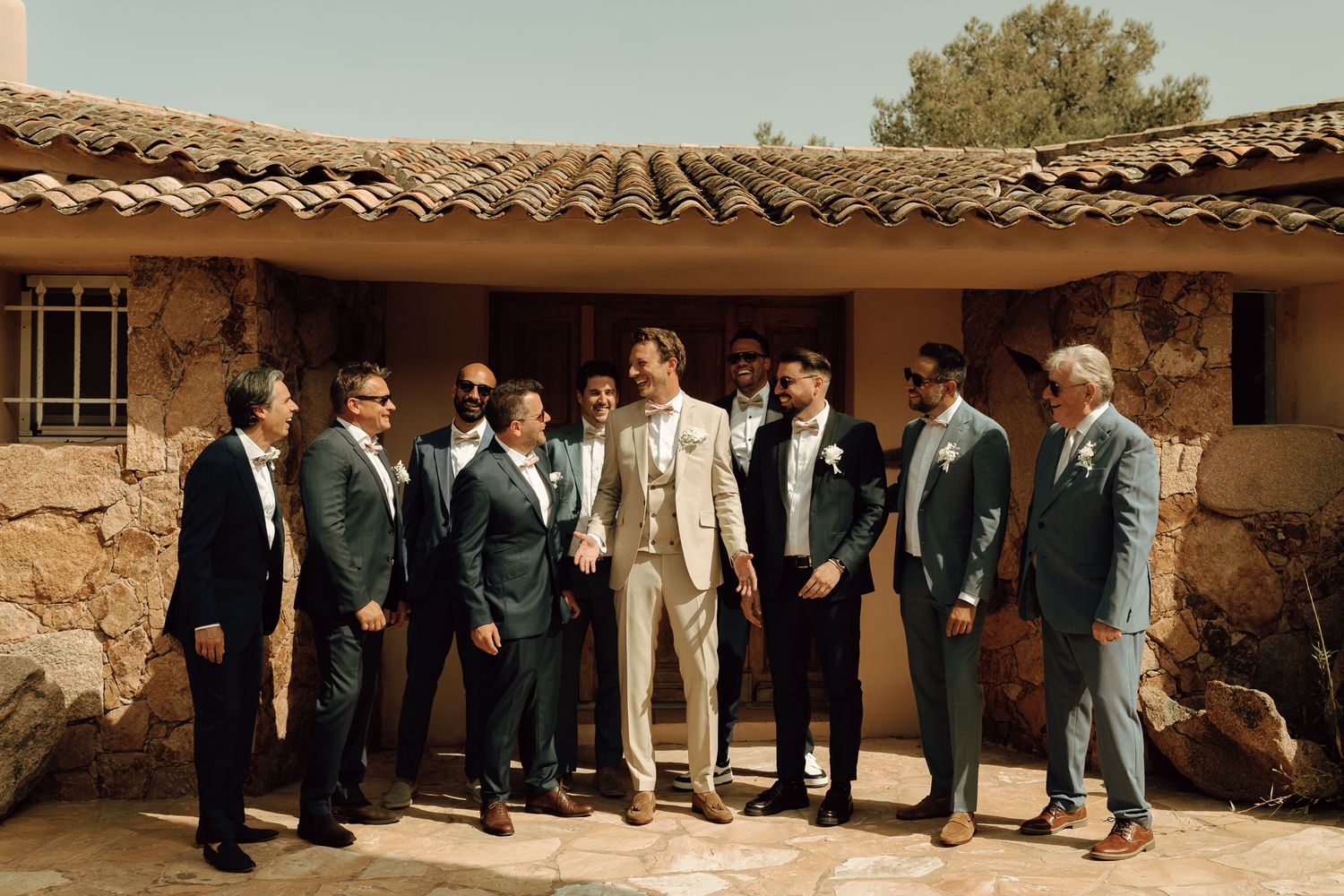 Groom in beige suit poses with groomsmen in dark suits outside a rustic stone wedding venue.