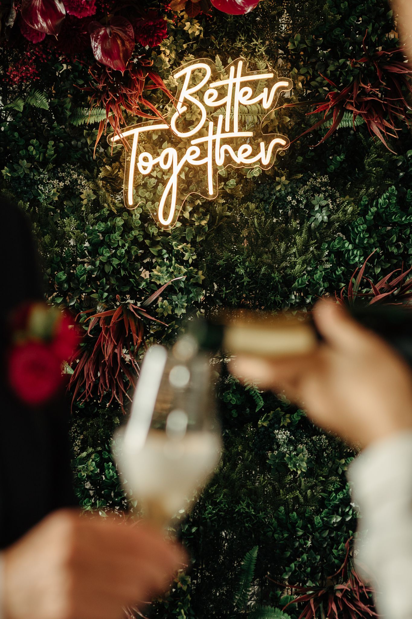 Neon 'Better Together' sign on lush green floral wall with champagne glasses in foreground at wedding reception.
