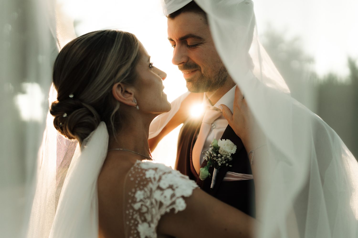 Bride and groom share an intimate golden hour moment wrapped together under a flowing white veil.