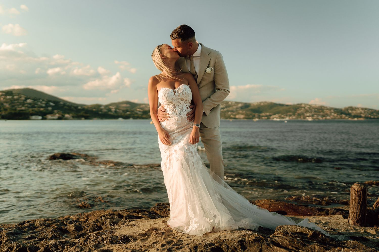 Bride and groom embracing tenderly on rocky seaside shore with scenic hills in the background.