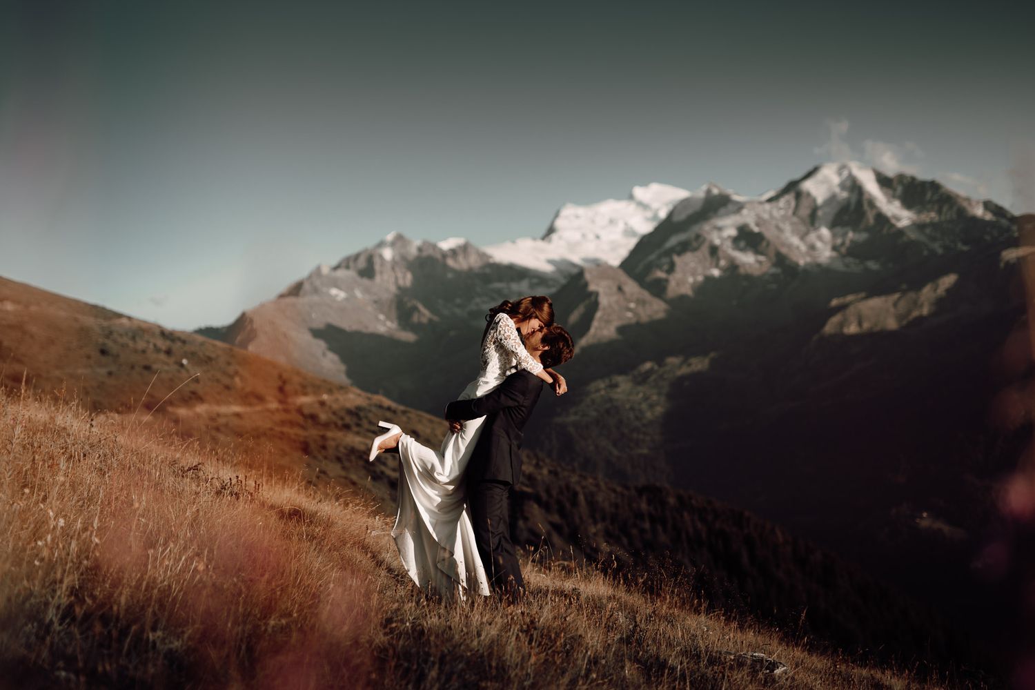 Couple embracing on a mountain hillside during their wedding, with snow-capped peaks in the background.