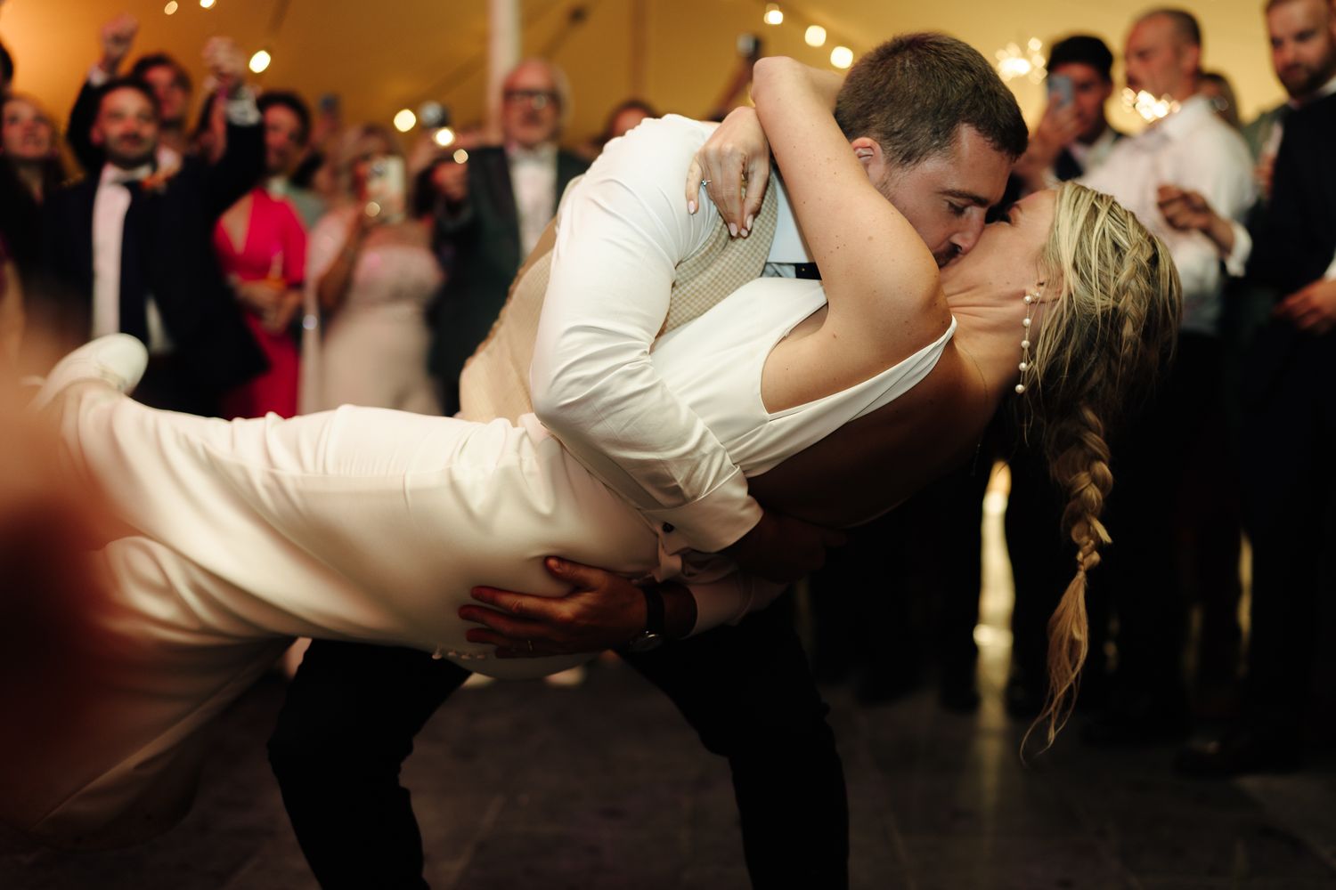 Groom dips bride during romantic first dance at wedding reception surrounded by guests.
