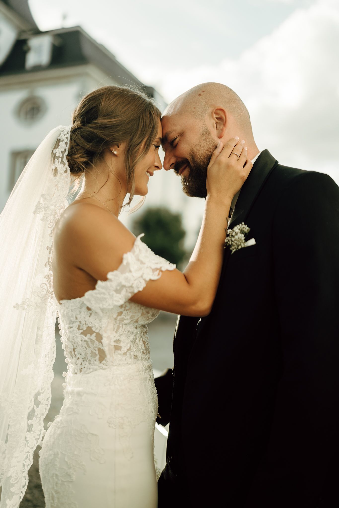 Bride and groom touch foreheads and smile intimately in front of a elegant chateau on their wedding day.