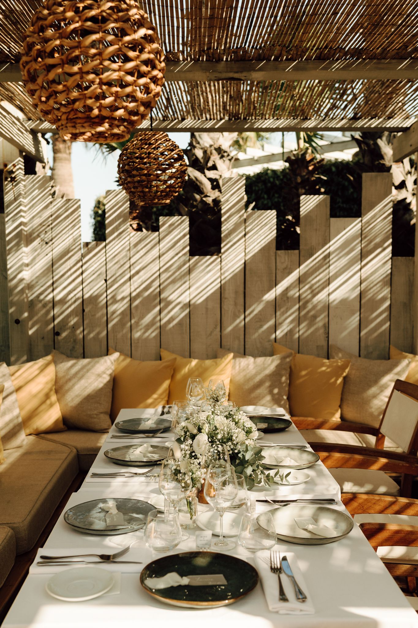 Rustic outdoor wedding reception table set with white florals under woven pendant lights and bamboo pergola.