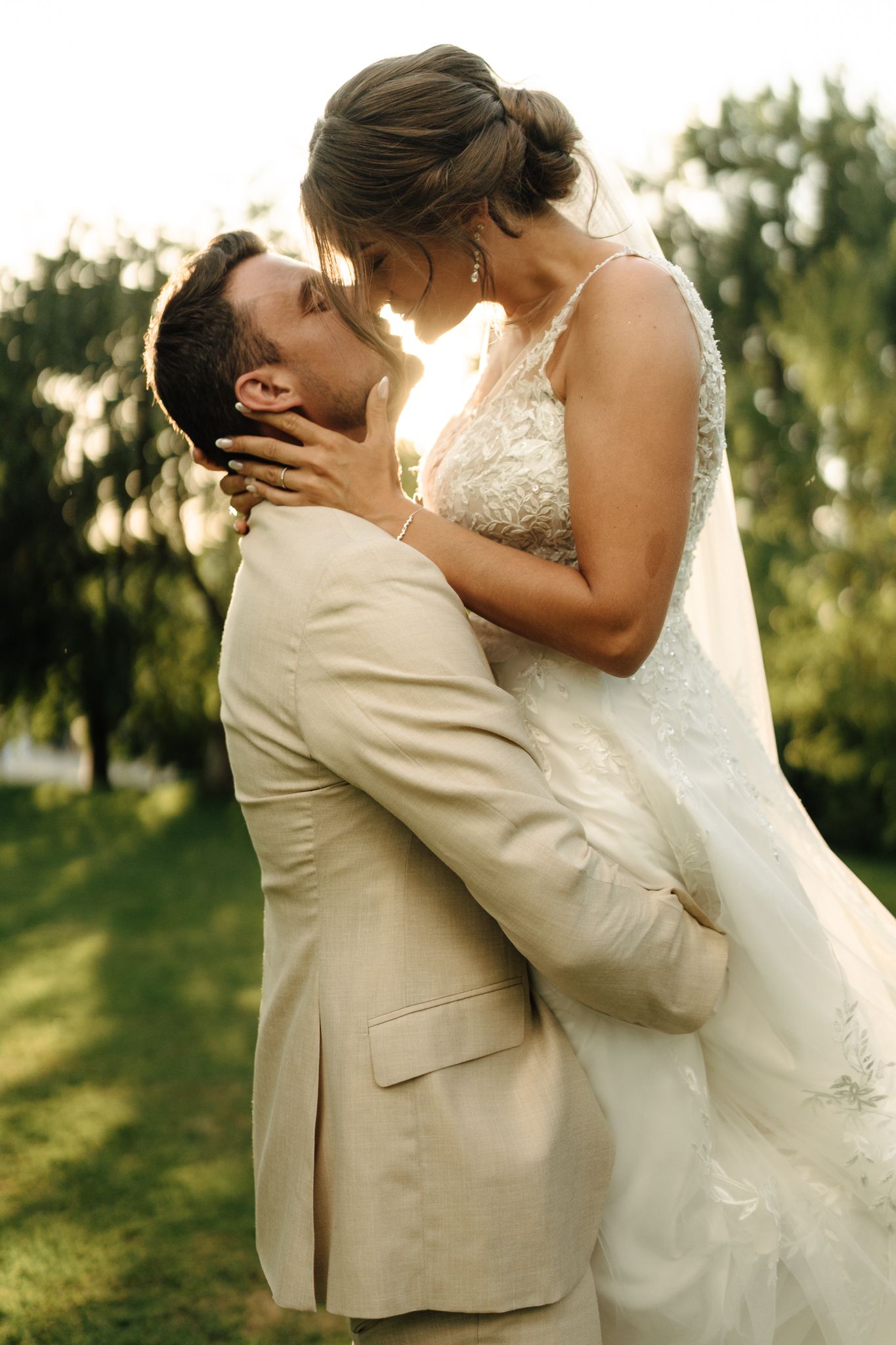 Groom lifting bride in lace wedding dress for a romantic sunset kiss in lush green park.