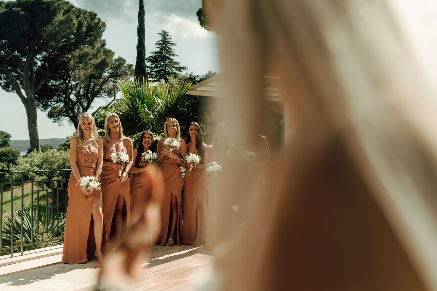 Bridesmaids in terracotta satin dresses holding white bouquets stand outside a sunny venue.