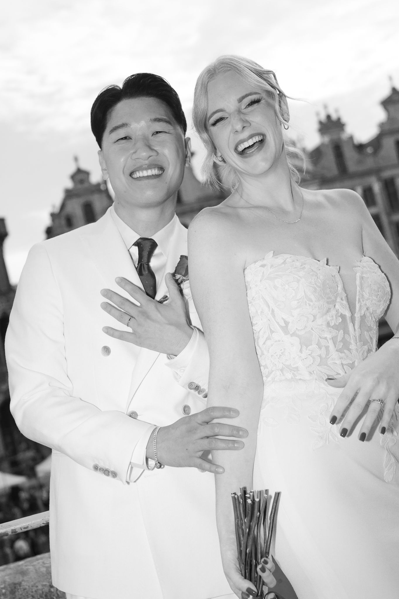 Black and white photo of a joyful interracial couple in wedding attire, laughing together outdoors.