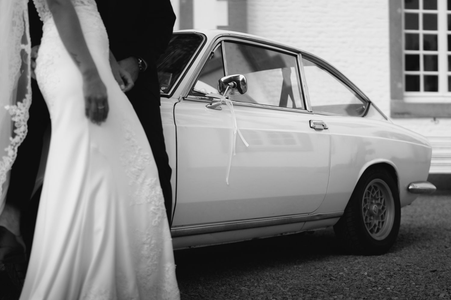 Black and white photo of bride in fitted lace gown standing beside vintage white classic wedding car.