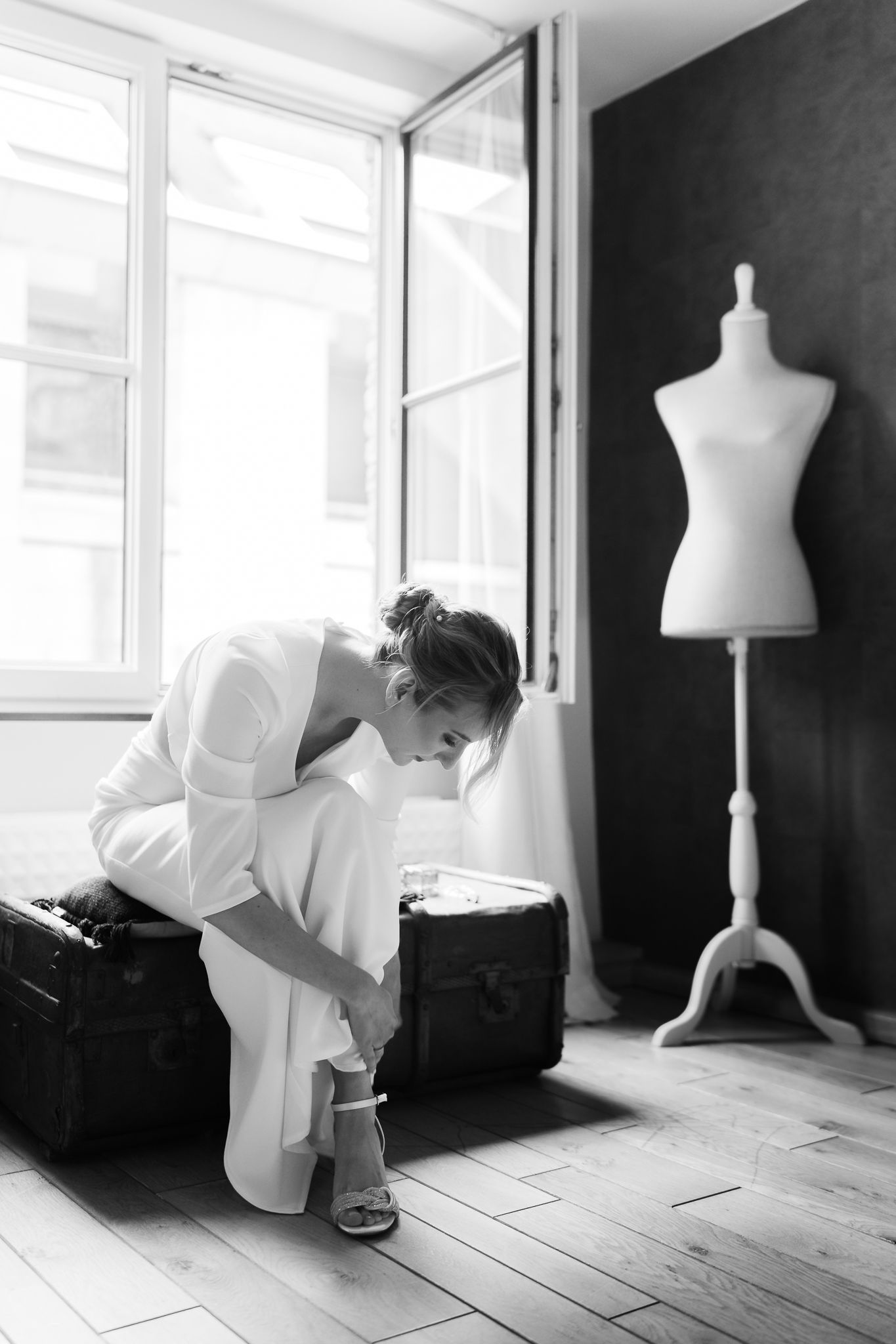 Black and white photo of bride putting on heels near vintage suitcase.
