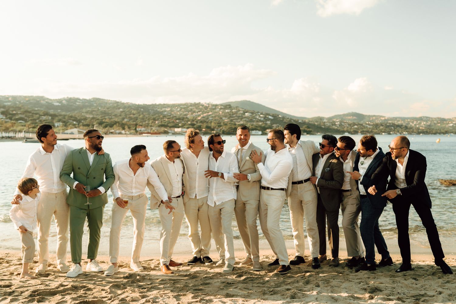 Group of groomsmen and male wedding guests laugh and pose together casually on a sunny sandy beach.