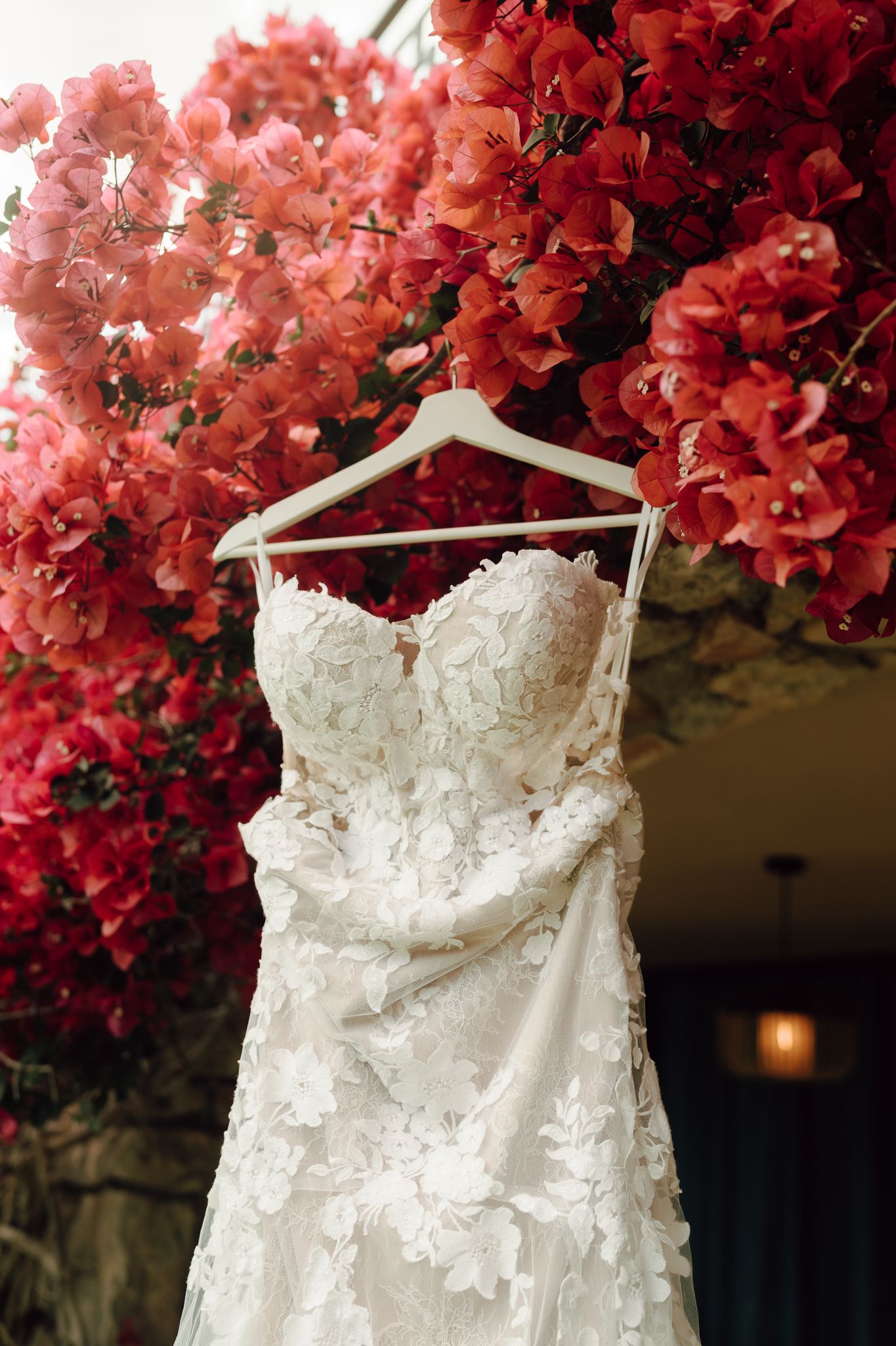Strapless floral lace mermaid wedding dress hanging on white hanger against vibrant red bougainvillea.