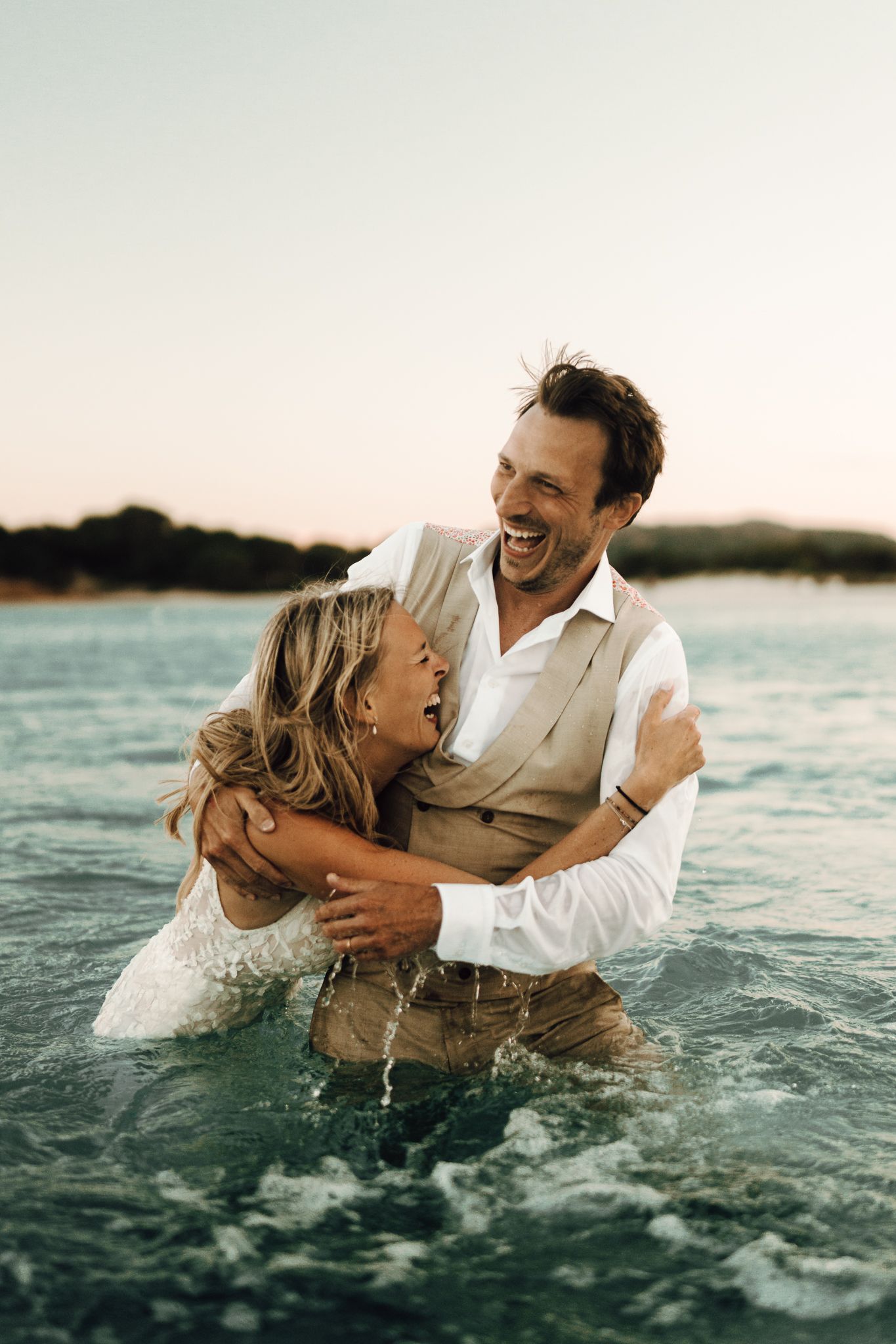 Laughing bride and groom embracing in ocean waves during beach wedding celebration.