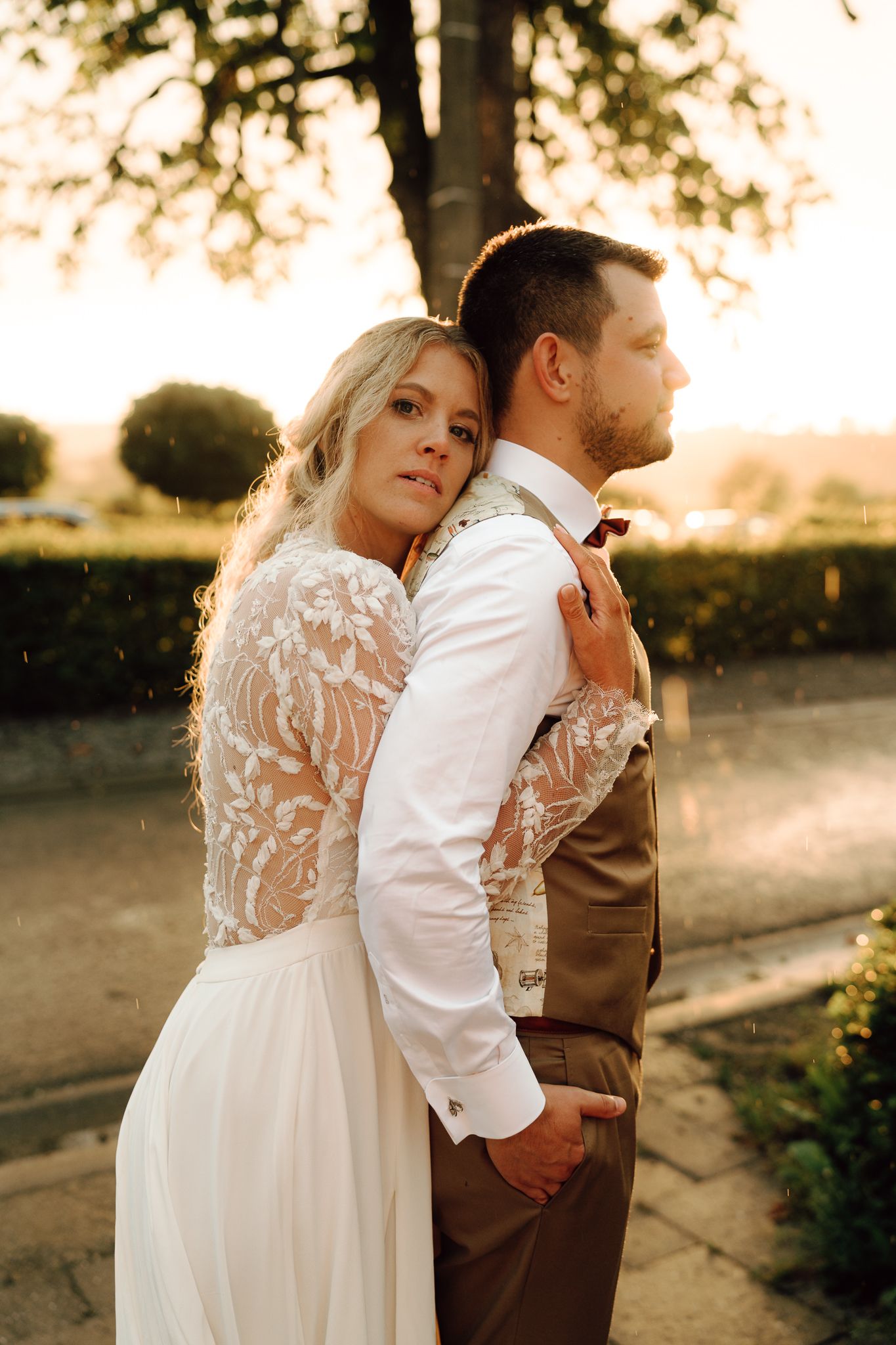 Bride in lace wedding dress embracing groom from behind during golden hour outdoors.