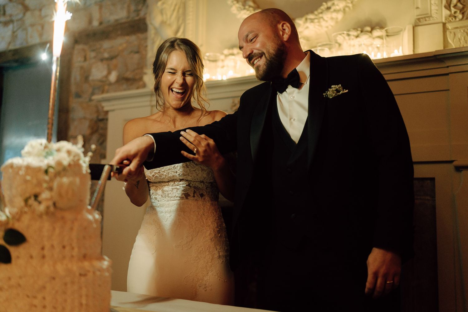 Joyful bride and groom laughing together while cutting their elegant tiered wedding cake at reception.