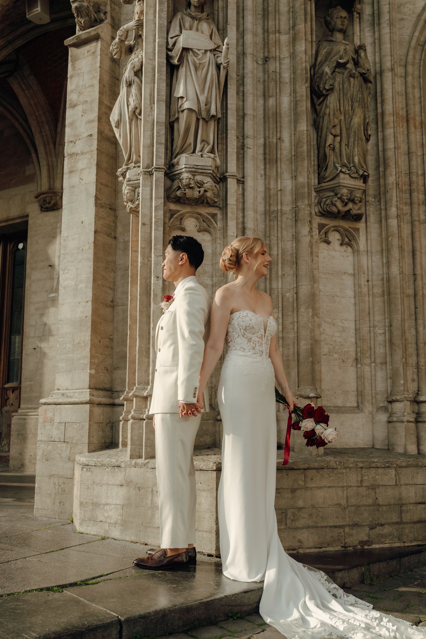 Bride and groom stand back-to-back in white wedding attire before ornate Gothic church statues, bride holding red bouquet.